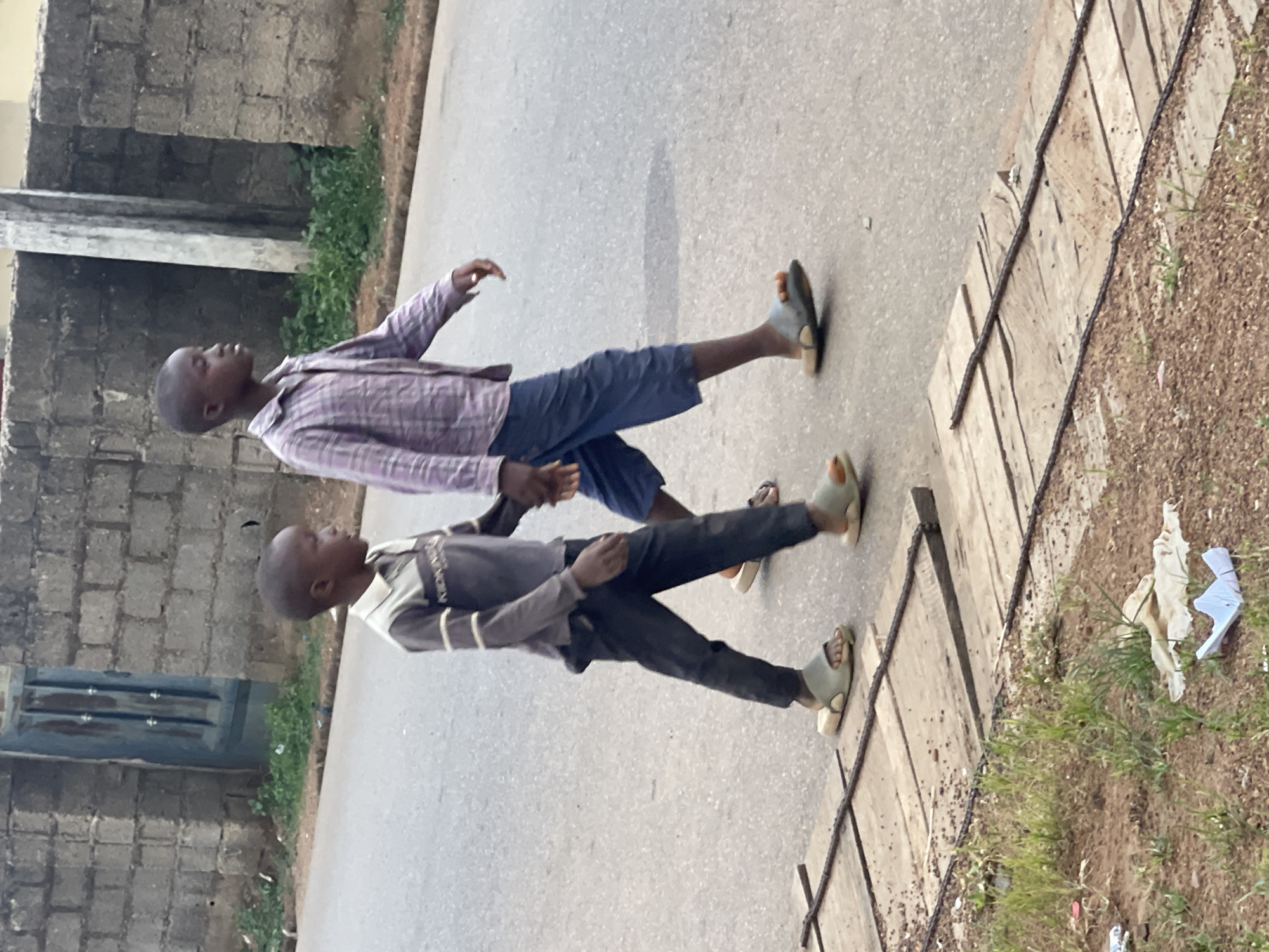 Two young boys are walking on a paved road in Akwanga, Nigeria. The boy closest to the camera wears a gray shirt with white stripes on the sleeves and dark pants. The other boy wears a plaid shirt and blue shorts. Both boys wear sandals. A concrete wall and greenery border one side of the road. On the other side is a wooden structure, possibly a drainage cover, next to an area with grass and some litter. The overall environment appears to be residential.