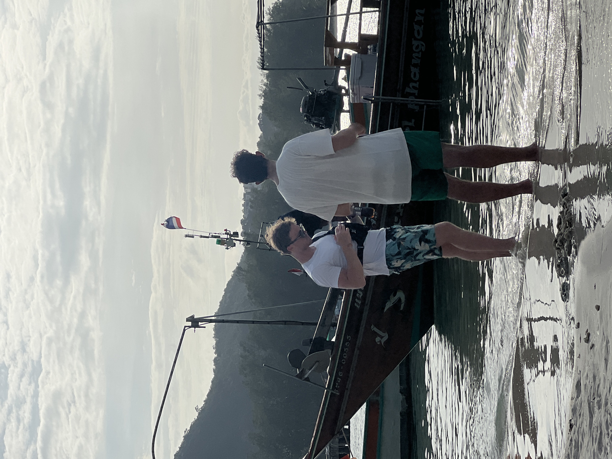 Two young adult males stand at the edge of a body of water, with their backs to the camera. The male on the left is wearing a white t-shirt, printed shorts, and a chest bag. He is looking down at his phone, which he is holding in his right hand. The male on the right is wearing a white t-shirt and green shorts. He is looking in the same direction as the other male. Behind the men are several boats docked in the water. One boat has a Thai flag flying from its stern. The water is calm and reflects the light. The background shows a lush green hillside. The sky is overcast with patches of sunlight breaking through. The scene appears to be a beach or harbor.