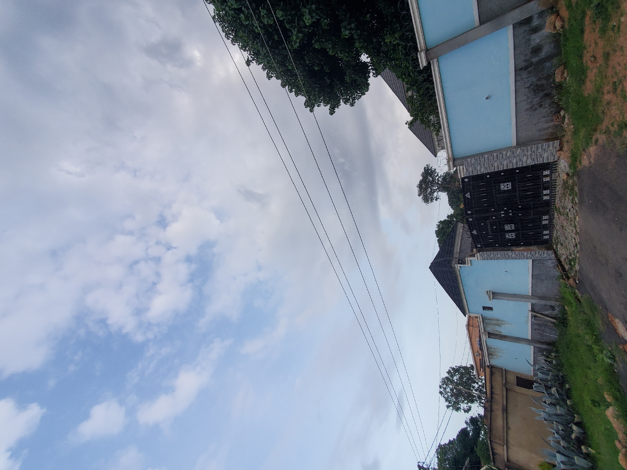 The image depicts a view of a residential property in Jos, Nigeria, characterized by a light blue stucco wall with grey concrete trim and a black metal gate with decorative patterns. The gate is set within a stone-clad entranceway. Above the wall, several power lines are visible against a sky filled with both bright white and darker grey clouds. To the right of the gate, a large, leafy green tree overhangs the property. In front of the blue wall, a section of a tan-colored wall is visible, with large agave plants growing in the foreground alongside patchy green grass. Further to the left, another section of the property with a tan wall and a dark doorway is seen. The ground in front of the gate is paved with cobblestones, transitioning to a dark asphalt road. The overall impression is a well-maintained residential entrance in a setting that appears to be undergoing some development or is in a tropical climate, judging by the vegetation.