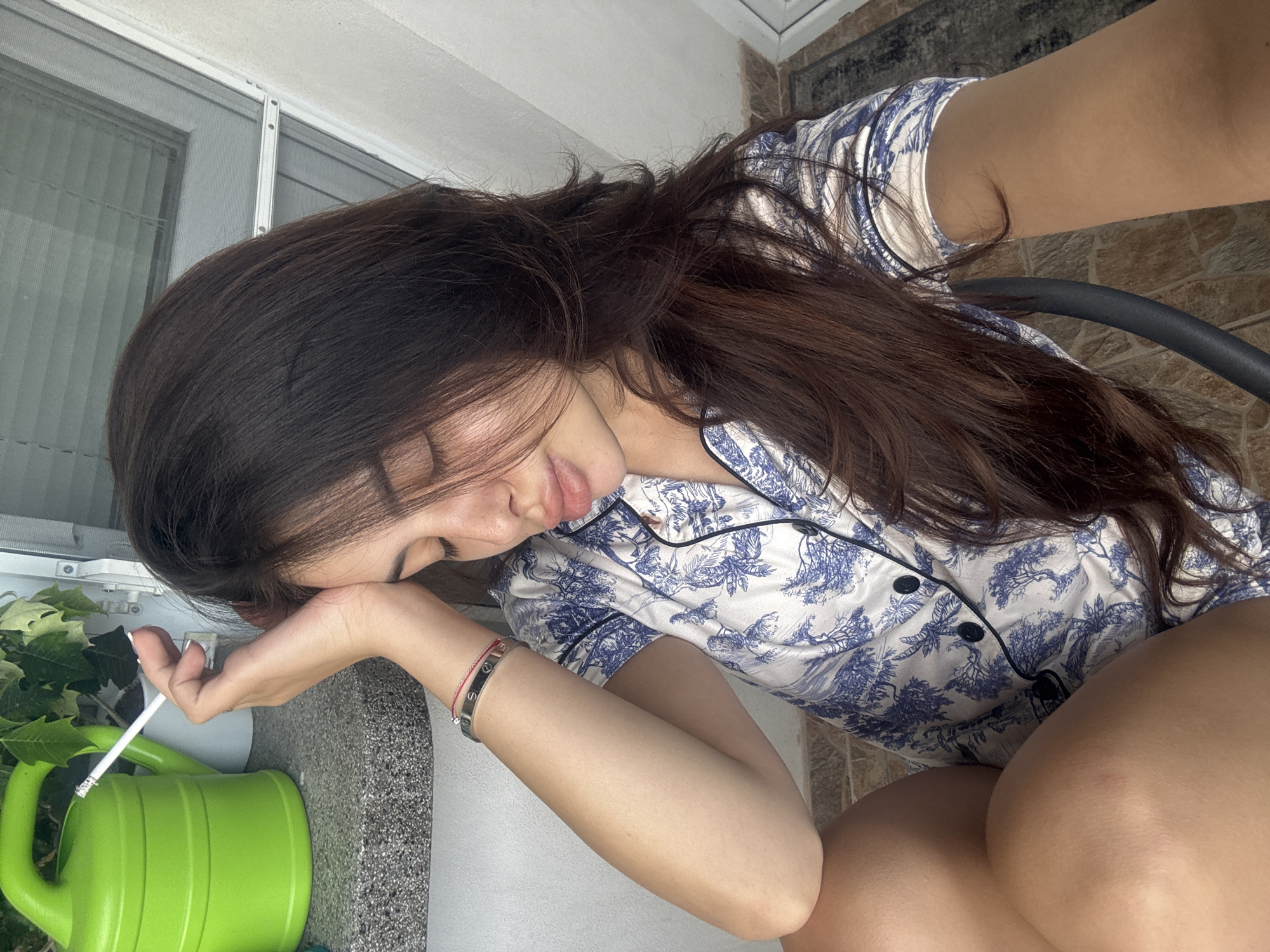 A young woman is pictured in a selfie taken outdoors. She is leaning her head on her right hand, with her eyes closed and lips slightly parted. She is holding a cigarette in her right hand. She is wearing a blue and white patterned pajama top. To her left, a green watering can and a potted plant are visible. The background shows a tiled floor and a window with blinds. The scene does not provide any specific indicators of the location being Plovdiv, Bulgaria.