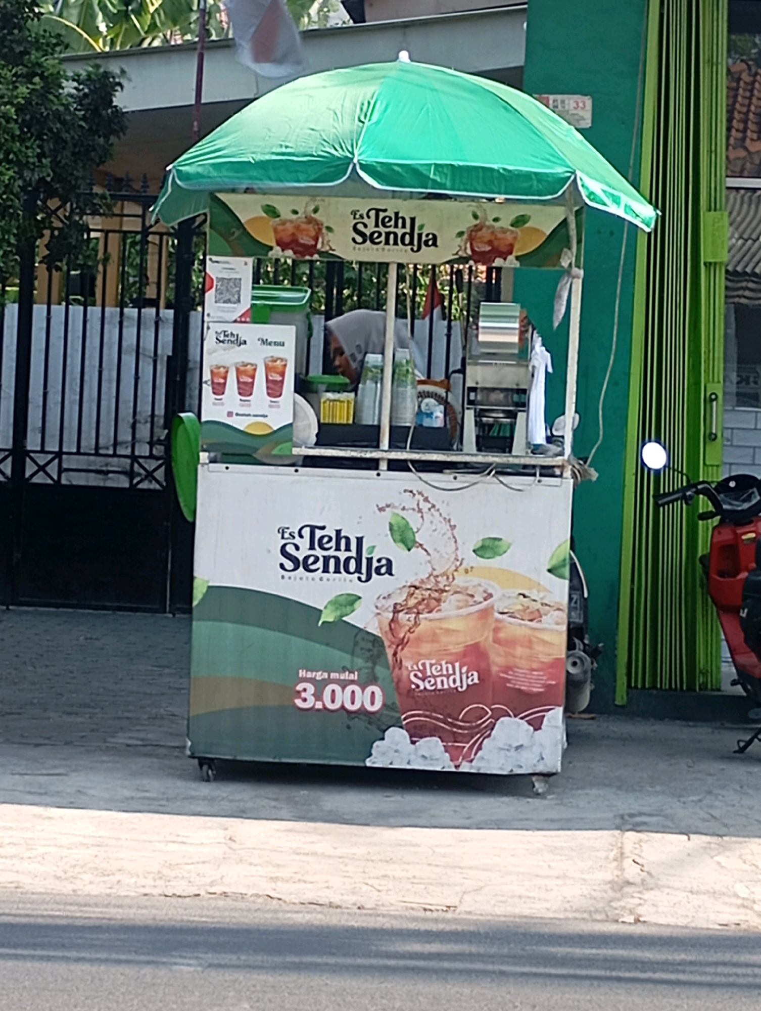 The image captures a street-level view of an "Es Teh Sendja" beverage cart under a green umbrella. The cart is positioned on a paved surface adjacent to a road, with a green metal gate and a building with green vertical slats in the background. A red motorcycle is partially visible to the right of the cart. The cart itself features branding for "Es Teh Sendja" with images of iced tea and the price "3.000". A person wearing a hijab is visible behind the counter, preparing drinks. QR codes and menu options are also displayed on the cart. The environment suggests a commercial area in Babelan, Indonesia, likely a roadside stall.