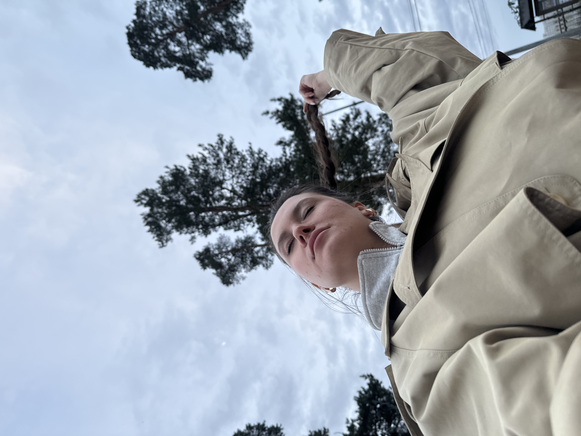 The image, originally rotated 90 degrees clockwise, presents an extreme low-angle outdoor shot of a person looking upwards. The subject, likely female, is positioned in the lower and central part of the frame, with her head tilted back and eyes closed, conveying a sense of calm or introspection. She is wearing a light beige or khaki trench coat over a grey hooded sweatshirt. Her dark hair, which appears to be styled in a braid or ponytail, is held up by her right hand, visible above her head. Small golden hoop earrings are visible on her ears.
The background is dominated by an overcast sky with patches of grey clouds. Dark green coniferous trees, resembling pines, are silhouetted against the sky, visible in the upper and lower sections of the frame. The diffused light indicates an overcast daytime. No direct sunlight is visible. The setting suggests a park or a natural area within an urban environment, consistent with locations in Moscow, Russia. There is no visible text in the image.