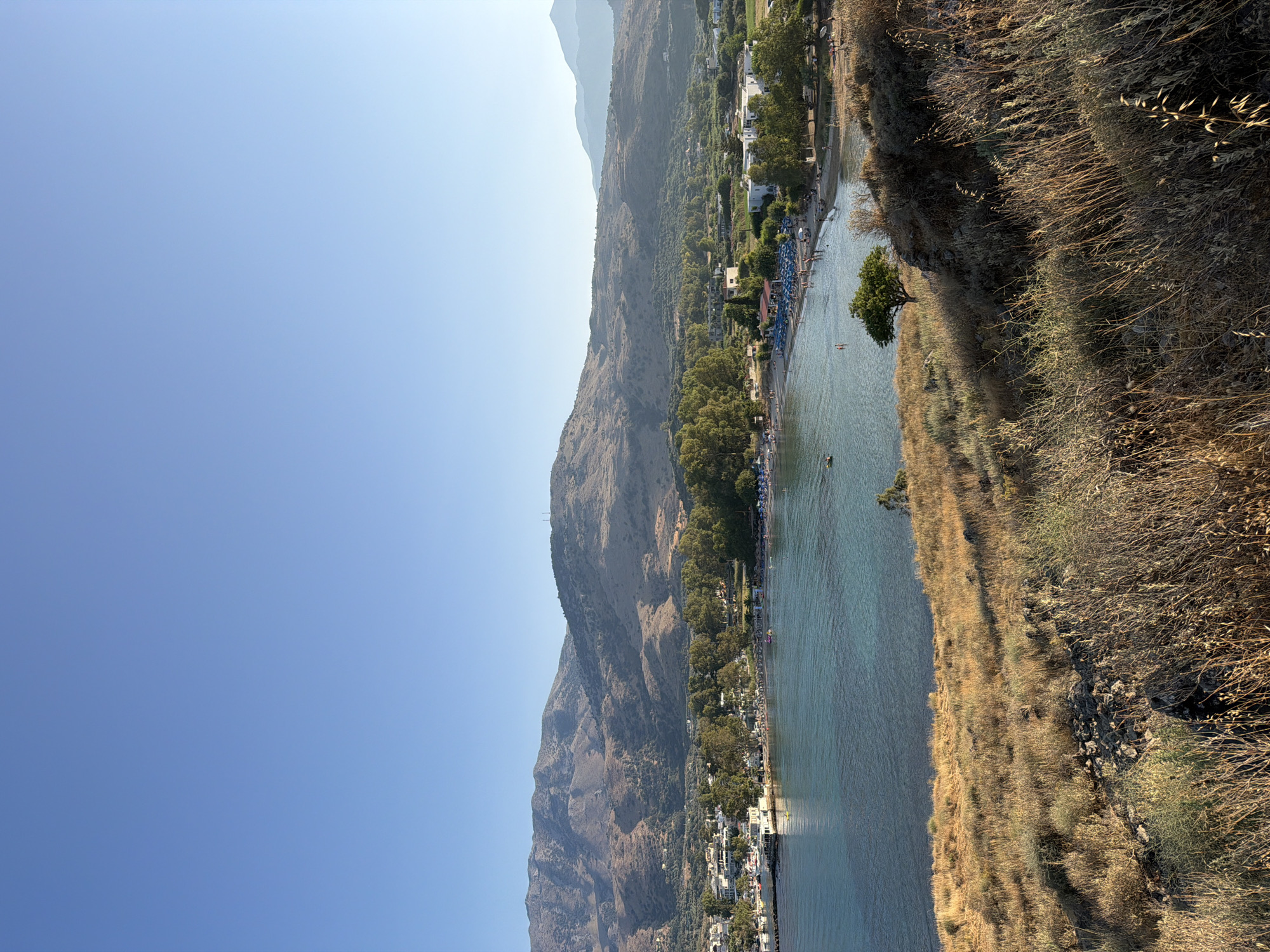 A bright, sunlit, medium-shot vertical image shows a panoramic view of Georgioupoli, Greece, on the island of Crete. The scene looks down and across a clear, turquoise bay. In the foreground, a dry, scrubby hill is covered in tall, golden-brown grasses and scattered rocks. A lone, small green tree stands on the hillside overlooking the bay.
The bay itself is tranquil, with a few small boats docked near a town on the left side of the image. A crescent-shaped beach lines the bay, dotted with numerous blue umbrellas and sunbathers. The water is calm and shallow near the shore, with a few people swimming.
Beyond the beach, the town of Georgioupoli is visible, with a cluster of white buildings and a few taller structures. Further inland, dense green trees and olive groves cover the lower slopes of rolling, barren mountains. The mountains dominate the background, their dusty, dry slopes catching the sunlight. The sky is a clear, bright blue with no clouds. The overall impression is one of a peaceful, sunny day in a coastal Greek town.