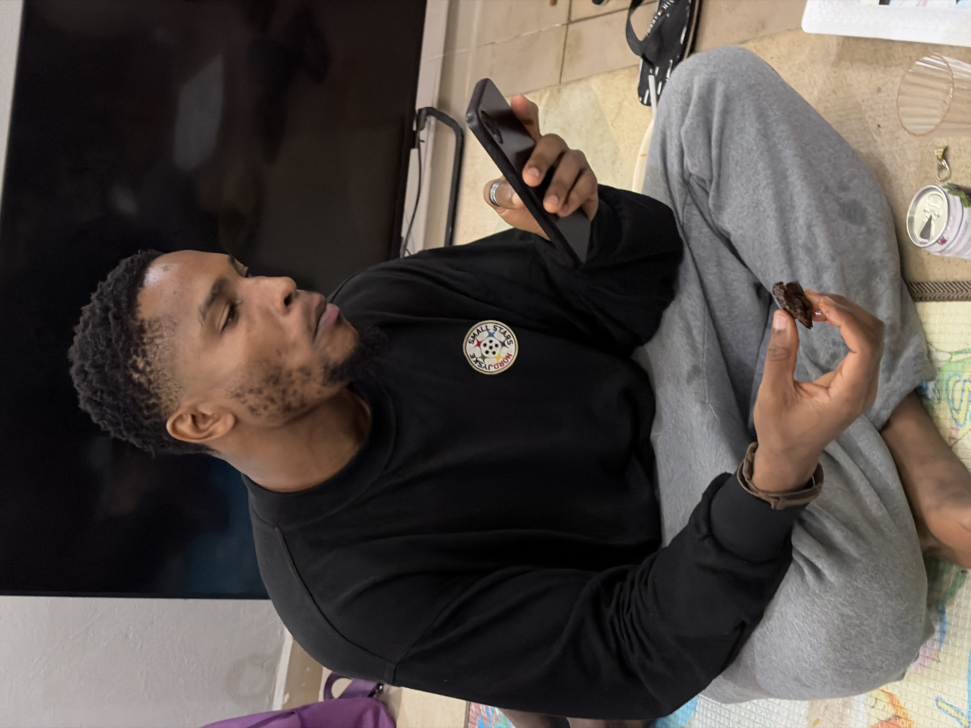 A man sits cross-legged on a patterned surface. He is holding a mobile phone in one hand and a small, dark piece of food in the other. He wears a black long-sleeved shirt with a circular emblem on the chest and grey sweatpants. A television sits in the background. A can, a clear glass, and a pair of flip-flops are nearby. There is no information available from the image to confirm the location in the city of Jos, Nigeria.