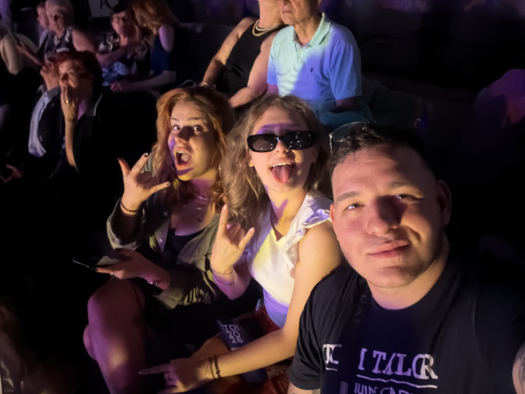 In a dimly lit indoor venue, possibly a concert hall or arena, a group of people are seated. The foreground is dominated by a selfie taken by a man with short dark hair, wearing a black t-shirt with white lettering. To his right, a woman with long blonde hair and sunglasses is sticking out her tongue. To her left, a woman with curly red hair and purple and yellow face paint is making a rock-on hand gesture and has her mouth open. Behind them, other individuals are visible, some with red hair and others with darker hair. One person in the background is wearing a light blue polo shirt. The seats appear to be dark colored. No specific landmarks in Plovdiv, Bulgaria, are identifiable within the frame of this image.