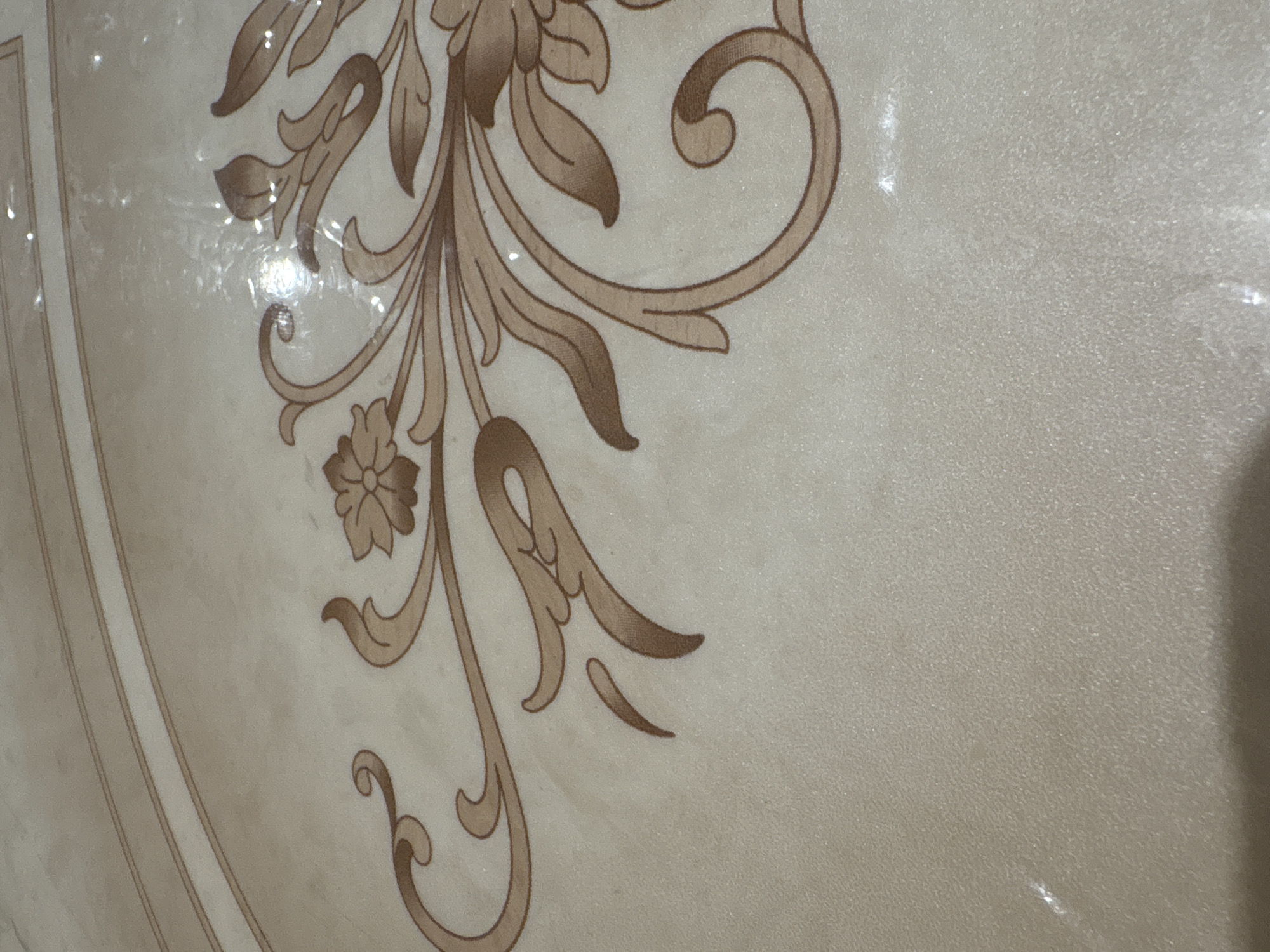 The image shows a close-up of a surface with a floral pattern. The pattern is primarily in shades of beige and brown, featuring stylized leaves, flowers, and curving tendrils. The background is a light beige color with a slightly textured appearance. The lighting suggests an indoor environment. The image doesn't provide sufficient detail to identify the location as Chaikovsky, Russia or provide any further specifics regarding people, subjects, objects, actions, interactions, or the environment.