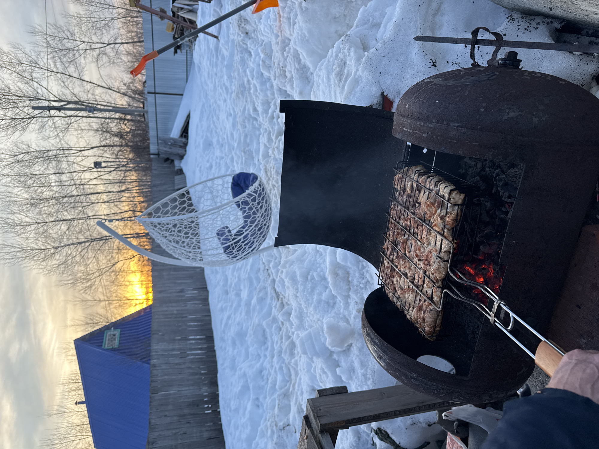 The image depicts an outdoor grilling scene at dusk in Chaikovsky, Russia, characterized by extensive snow cover on the ground. In the foreground, a person's left hand, clad in a dark sleeve, operates a metal grilling basket containing numerous pieces of raw or partially cooked meat over an active charcoal fire. The fire burns within a dark, repurposed metal barrel serving as a grill, from which smoke rises. To the immediate left, a portion of a rough wooden structure is visible. The midground features a snow-covered yard with uneven terrain. A white, egg-shaped wicker hanging chair with a visible blue cushion is suspended to the right of the grill. A weathered wooden fence horizontally spans the scene, separating the immediate area from the background. Beyond this fence, a house with a steep, bright blue corrugated metal roof and a small, decorative attic window is visible. The horizon behind the house displays a prominent orange glow, indicative of a sunset. Numerous bare deciduous trees and a utility pole are silhouetted against the sky in the distant background. To the far right, a section of a corrugated metal fence and an orange-handled shovel leaning against a support are present.