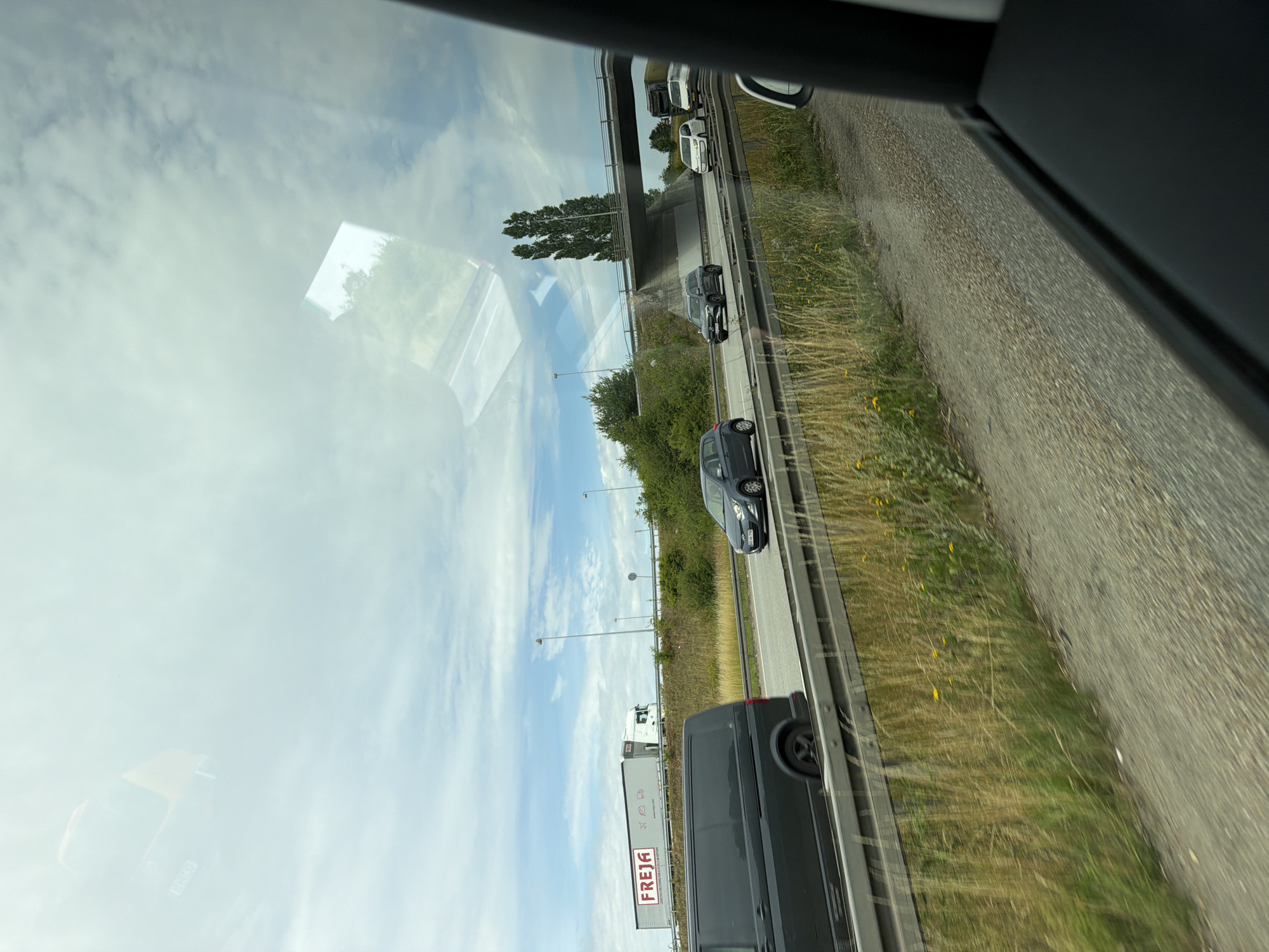 A highway scene is depicted from inside a vehicle. The focus is on the road ahead, with several vehicles visible. On the left, a dark grey van is in the foreground, moving on the highway. To its right, a dark grey sedan is also in motion. Further ahead, a white sedan and a light-colored van are visible. A large truck with a white trailer displaying the "FREJA" logo is in the distance on the left side of the highway. A bridge arches over the highway in the background, with tall trees visible behind it. The sky is partly cloudy with patches of blue. The surroundings include grassy areas and vegetation alongside the highway. The image appears to have been taken from a moving vehicle, suggesting a journey through the city of Karlslunde, Denmark.