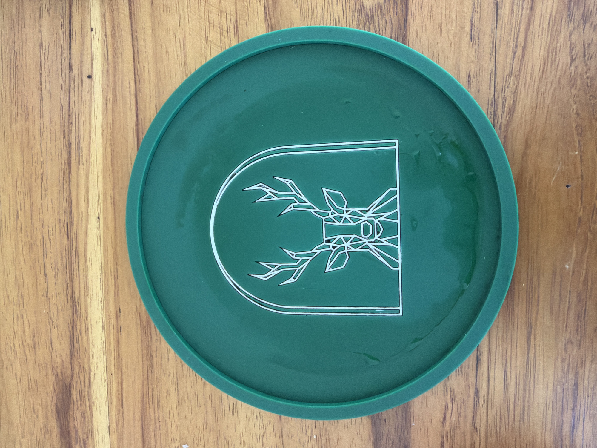 A green, circular coaster with a white outline of a deer's head inside an arch is placed on a wooden surface. The coaster is centrally located in the image. The wooden surface has a visible grain pattern. The image is taken from a top-down perspective. There is no indication of the location being Baturiti, Indonesia.
