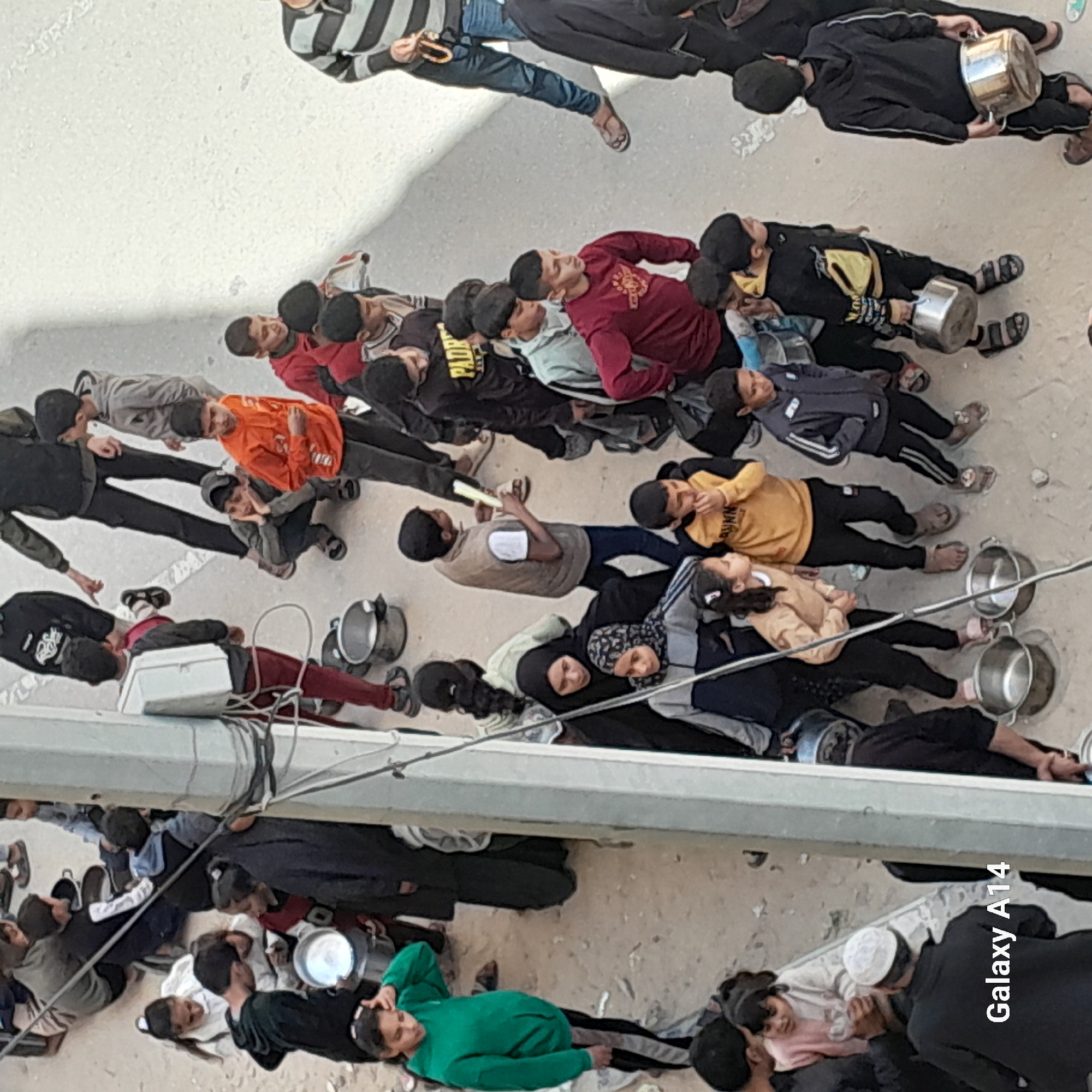 In the city of Unknown, Unknown, an aerial shot shows a large group of people, mostly children and some adults, lying on the ground. The individuals are primarily dressed in casual clothing. Many of the children are holding or have next to them what appear to be metal pots or containers. A few are carrying white boxes. Some are lying next to each other in pairs or small groups, while others are more isolated. An adult is seen standing and observing the scene. Overhead are pipes or beams, obscuring part of the view. The location appears to be a paved outdoor area.