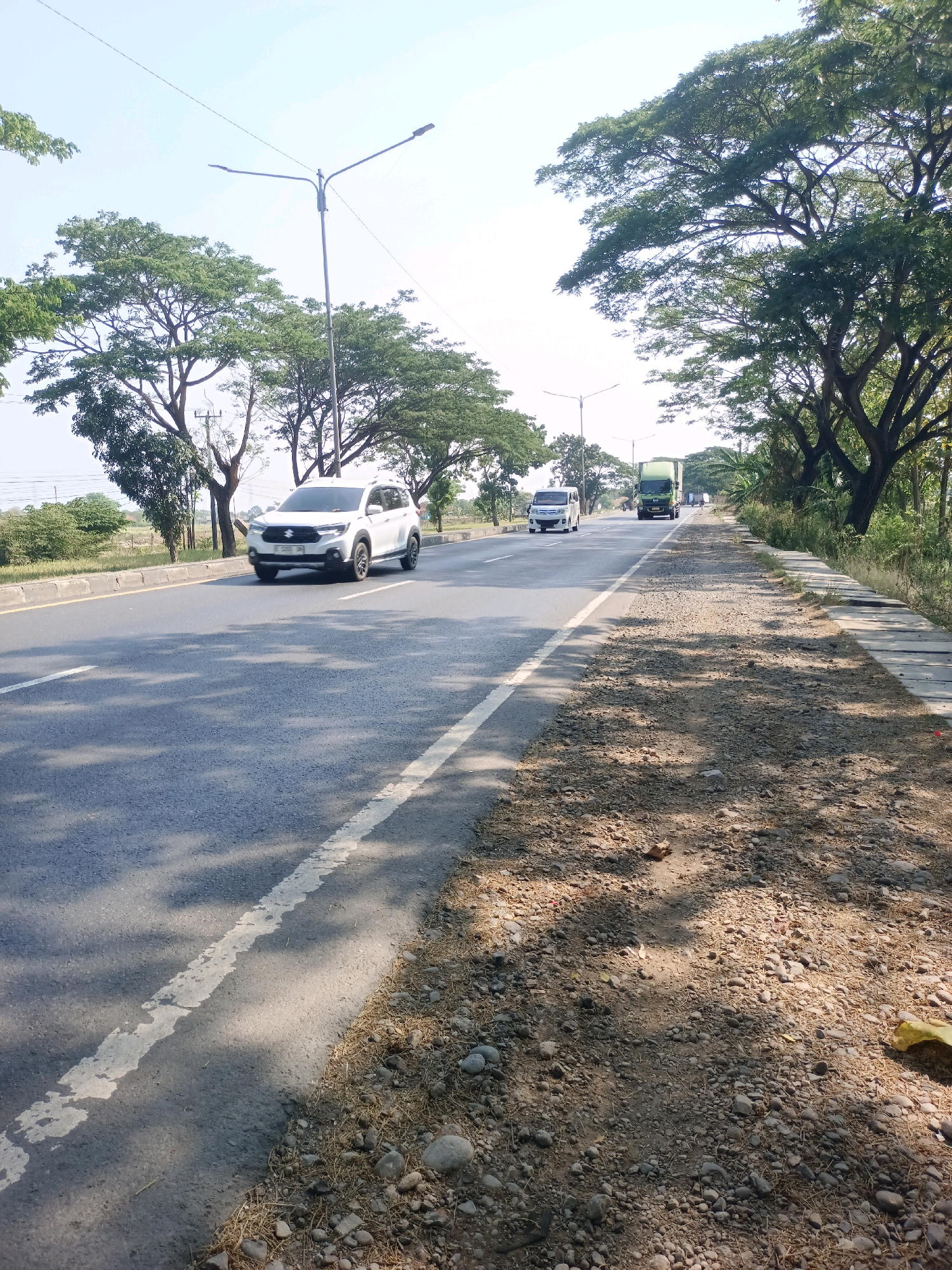 A low-angle, eye-level, wide shot captures a sunlit day along a two-lane highway in Subang, Indonesia. The asphalt road, marked with a white broken line, dominates the foreground and midground. A white SUV is traveling in the left lane, while further ahead, a white minivan and a green truck are also in motion. Lush green trees with expansive canopies line both sides of the road, casting dappled shadows across the tarmac. A gravel shoulder with scattered stones and dry grass borders the right side of the road, transitioning to a concrete drainage system. The sky is clear and pale blue. The scene is devoid of people.