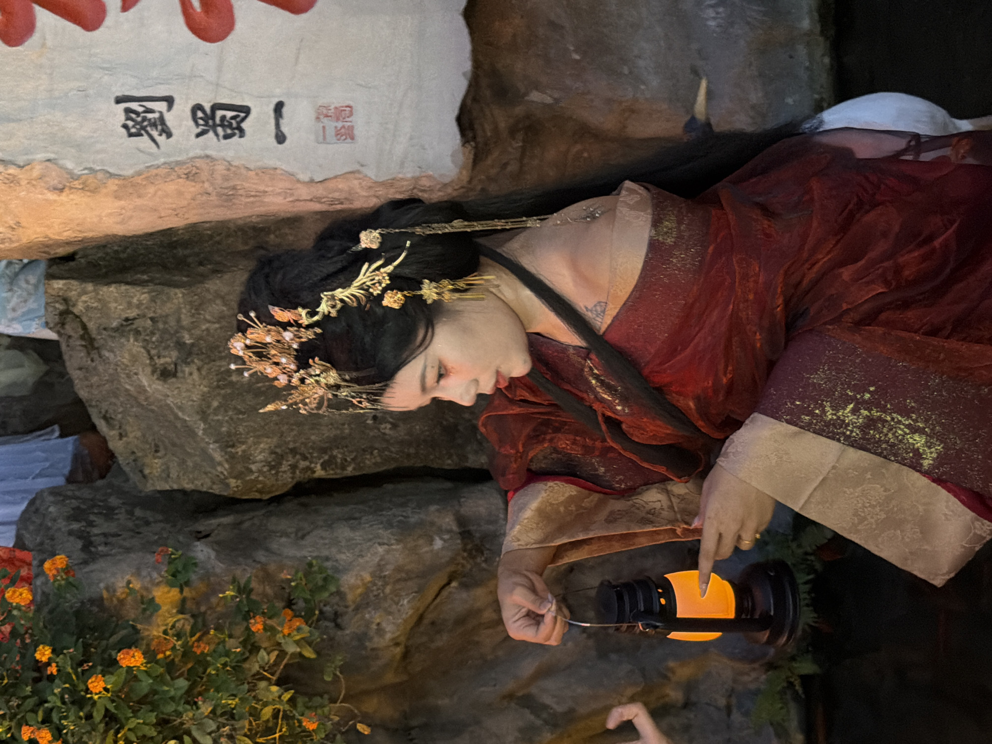 A woman dressed in traditional Chinese attire, with an elaborate gold headpiece adorned with pearls and dangling gold ornaments, holds a small, black lantern with an orange light emanating from it. She is positioned in front of a rocky outcrop and a light-colored scroll with Chinese calligraphy. Her red, gold-trimmed garment features shimmering gold embellishments. Her gaze is directed downwards and to her left, and her left index finger is extended, lightly touching the lantern. The background includes orange flowers on a bush to the left, and large, grey rocks. The image appears to have been taken in Yuzhong District, China.