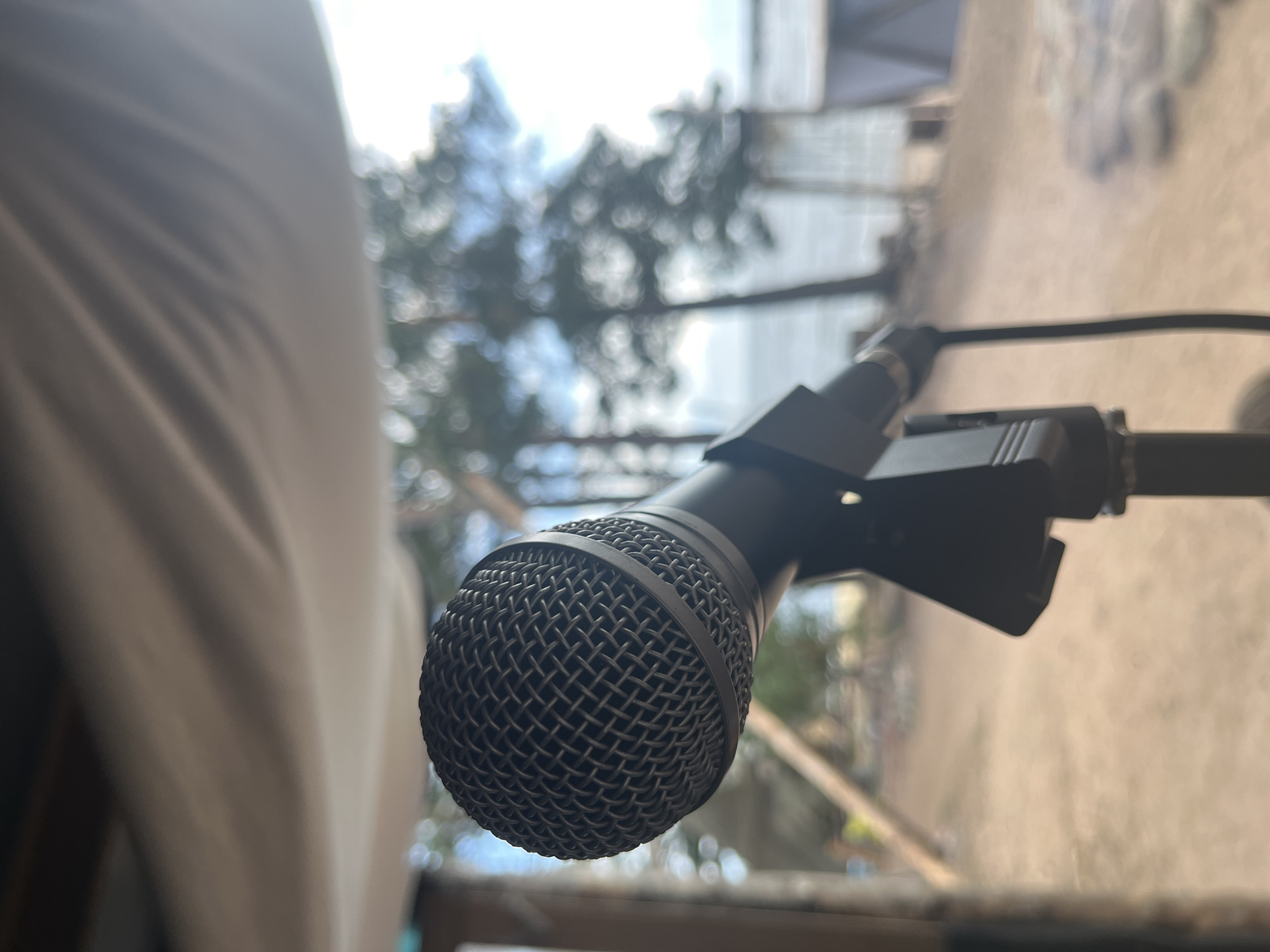 A microphone on a stand is in the foreground of an outdoor scene. The microphone is black and has a mesh grill. The stand is also black. Behind the microphone, out of focus, are trees, the sky, and what appears to be water. There is also a white tarp overhead. The location is described as Unknown, Unknown.
