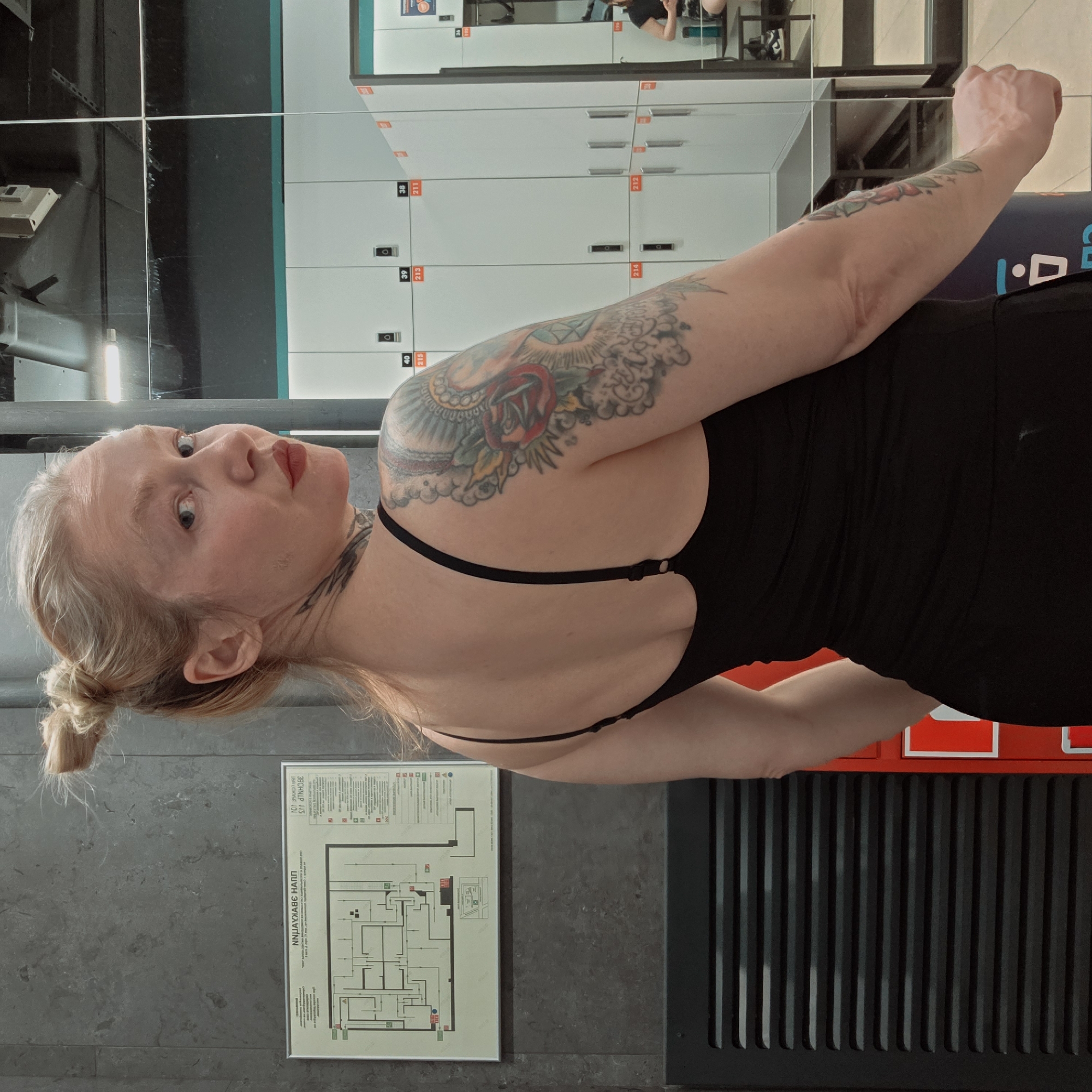 A woman with fair skin, blonde hair styled in a bun, and multiple tattoos on her arms and neck is posing for a photograph in what appears to be a locker room. She is wearing a black, backless top. Behind her are rows of lockers, and a framed emergency exit plan is mounted on the wall. Based on the wall-mounted evacuation diagram with text in Russian, the location is likely Saint Petersburg, Russia.