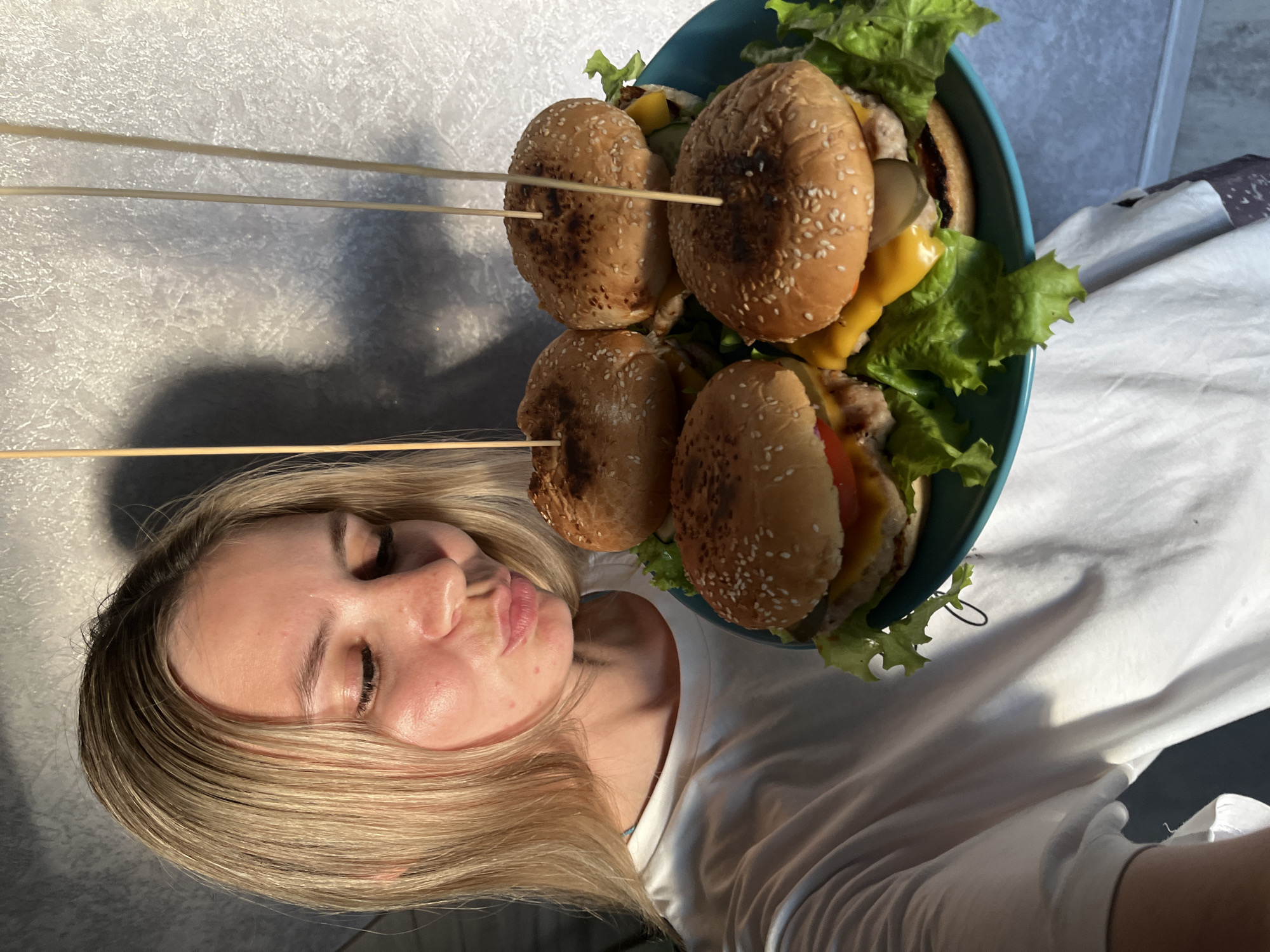 A young woman with blonde hair, wearing a white t-shirt, is taking a selfie while holding a blue plate with four hamburgers. The hamburgers are on sesame seed buns and topped with lettuce, cheese, pickles, and tomato. The background is a gray textured wall with a shadow of the woman and a few wooden skewers. The scene appears to be indoors. There is no information in the image to suggest the location is in Chaikovsky, Russia.