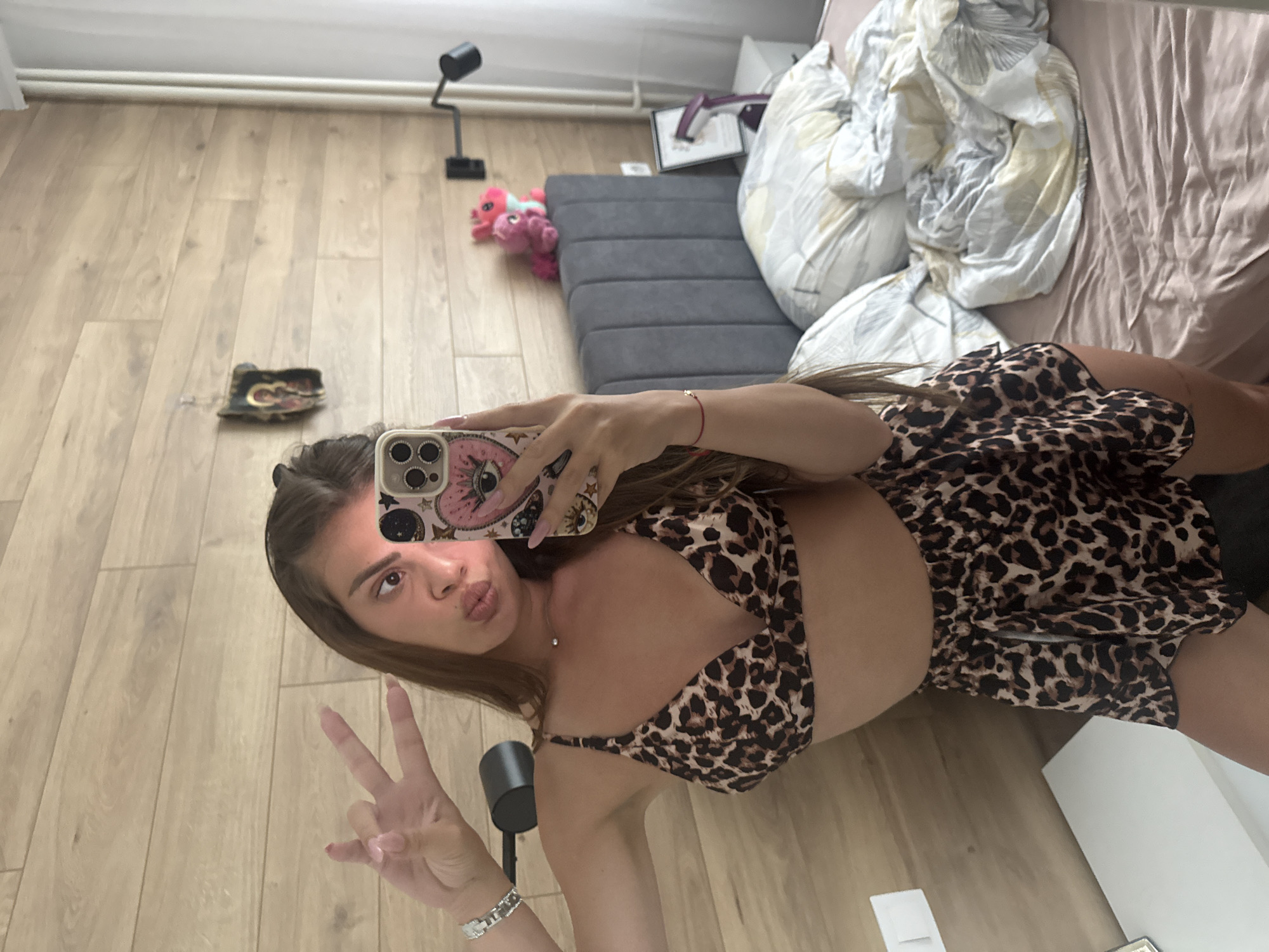 A mirror selfie shows a person, presumably female, wearing a leopard-print top and shorts, posing with a peace sign. The reflection shows a room interior featuring a wooden floor, a grey sofa, a bed with unmade sheets, and miscellaneous items such as a desk lamp and a stuffed animal. The city and specific location are unknown.