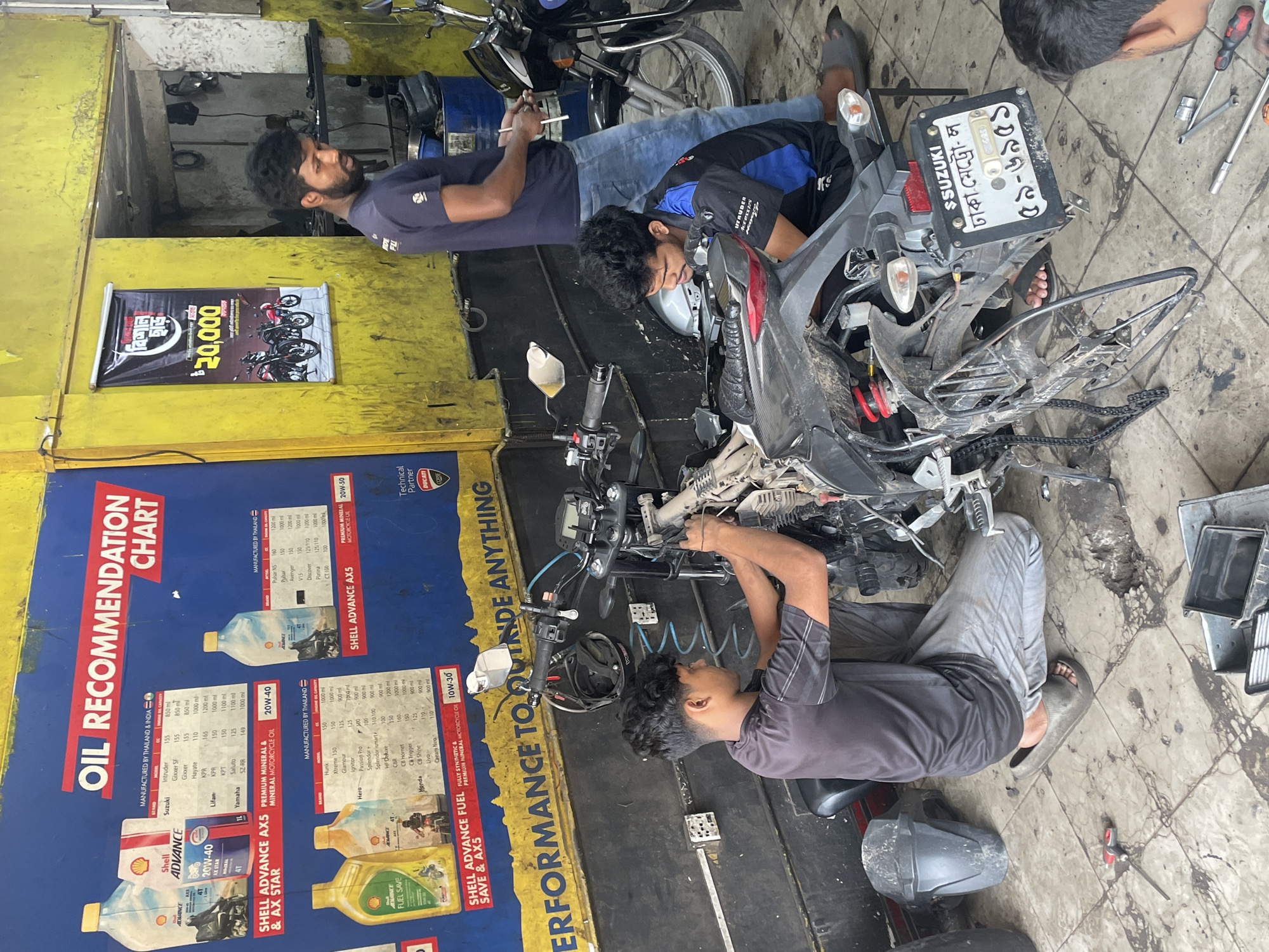 Three men are repairing a motorcycle in what appears to be a motorcycle repair shop. One man, wearing a navy blue shirt and jeans, is working on the motorcycle to the left. Another man in a blue and black shirt is inspecting a grey Suzuki motorcycle. The third man, wearing a grey shirt and grey pants, works on the motorcycle's front end. The motorcycle is partially disassembled, with parts scattered around. The environment is a garage setting, with an "OIL RECOMMENDATION CHART" displayed on the wall which includes the SUZUKI logo. The license plate on the Suzuki motorcycle indicates that the scene is in Dhaka, Bangladesh, specifically designated as "ঢাকা-মেট্রো-ল".