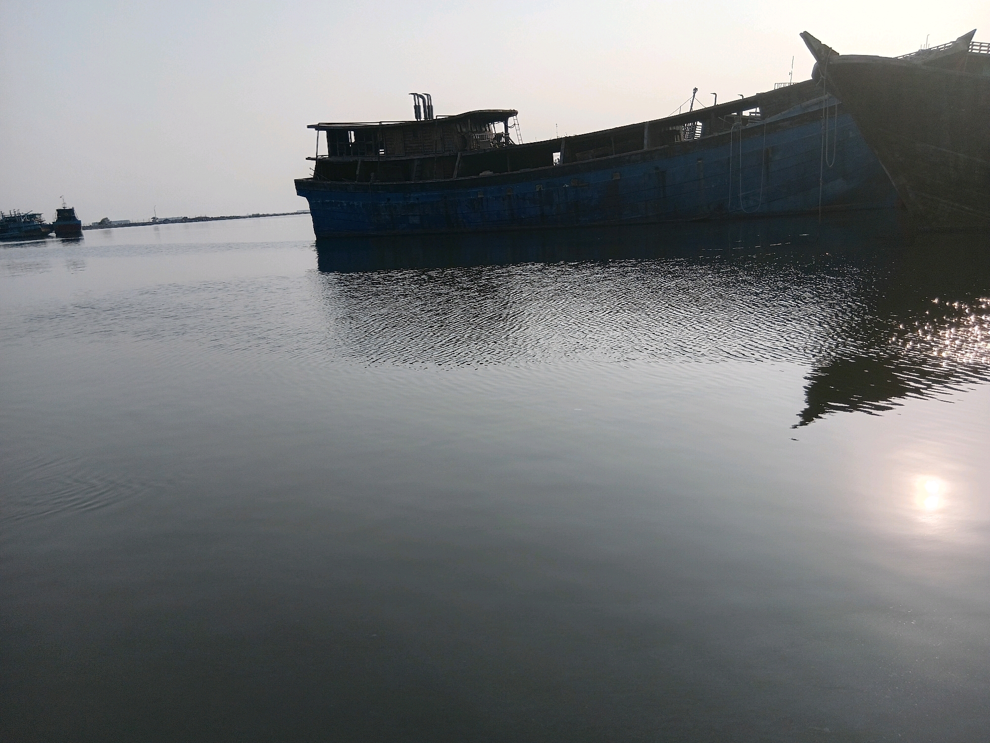 In Cirebon, Indonesia, two weathered, dark-hulled ships, possibly decommissioned or under repair, are anchored in a tranquil body of water under an overcast sky. The larger vessel, painted a faded blue and showing signs of disrepair with visible wood paneling and a partially constructed upper deck, dominates the frame, its reflection subtly rippling on the water's surface. A second, smaller ship with a red hull is visible in the distance to the left. The environment appears to be a harbor or a shipyard, with a faint, indistinct shoreline in the background suggesting industrial or urban proximity. The water is calm, with gentle waves and scattered reflections of the sky and ships, hinting at a quiet, possibly early morning or late afternoon scene.