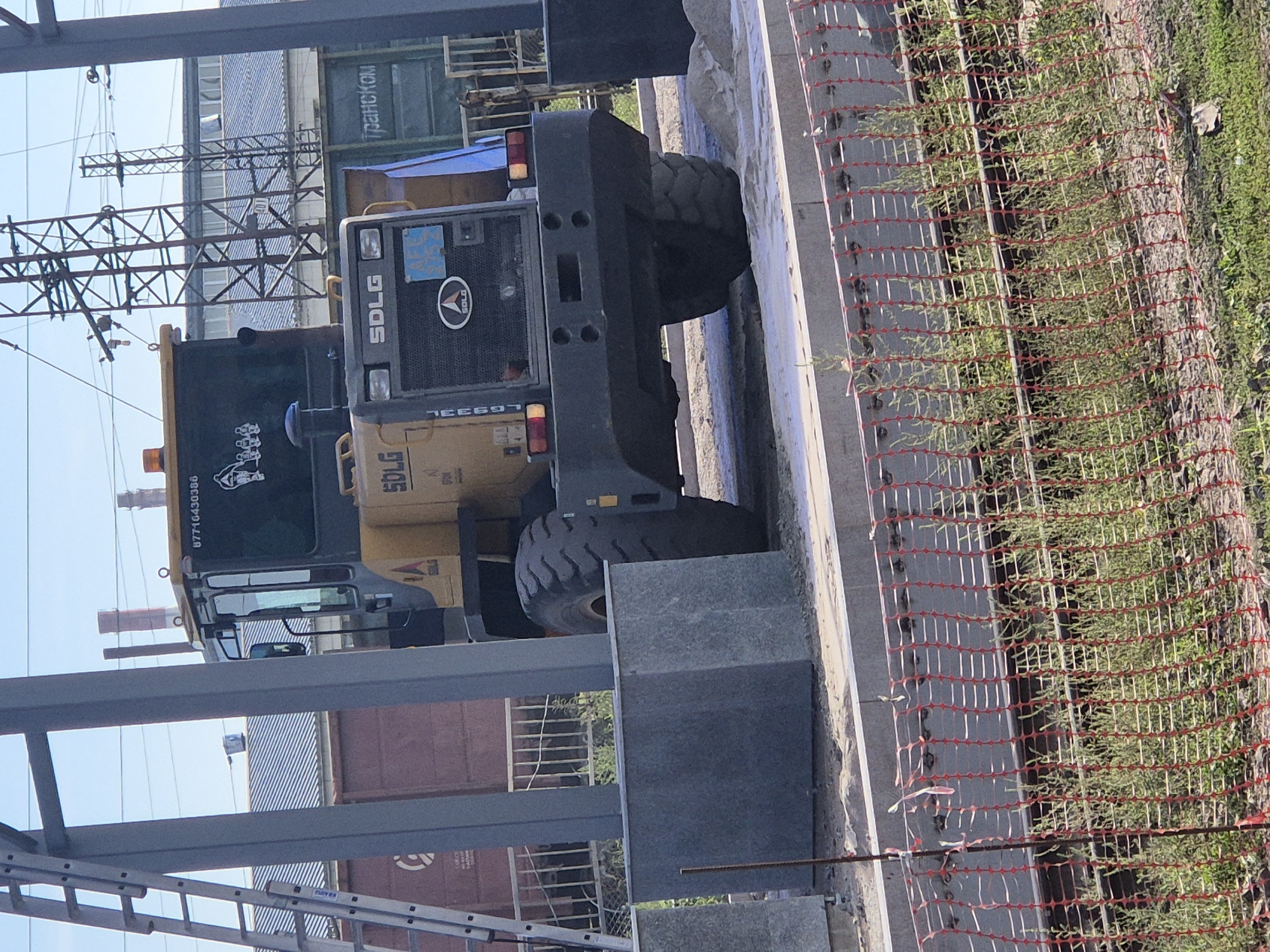 An outdoor, daytime, eye-level, medium shot captures the rear of a yellow SDLG wheel loader parked on a construction site in Astana, Kazakhstan. The loader's prominent black bumper, large tires, and rear lights are visible. Behind the loader, several elements of the urban environment are present: a railway car with Russian text "Транском" painted on its side, power lines supported by metal towers, and a partially constructed building with a blue tarp. A tall ladder leans against a grey pole on the left side of the frame. In the foreground, a red safety net divides the construction area from a patch of green vegetation growing between railway tracks. The scene suggests ongoing construction or infrastructure work in an industrial or transportation hub within Astana.