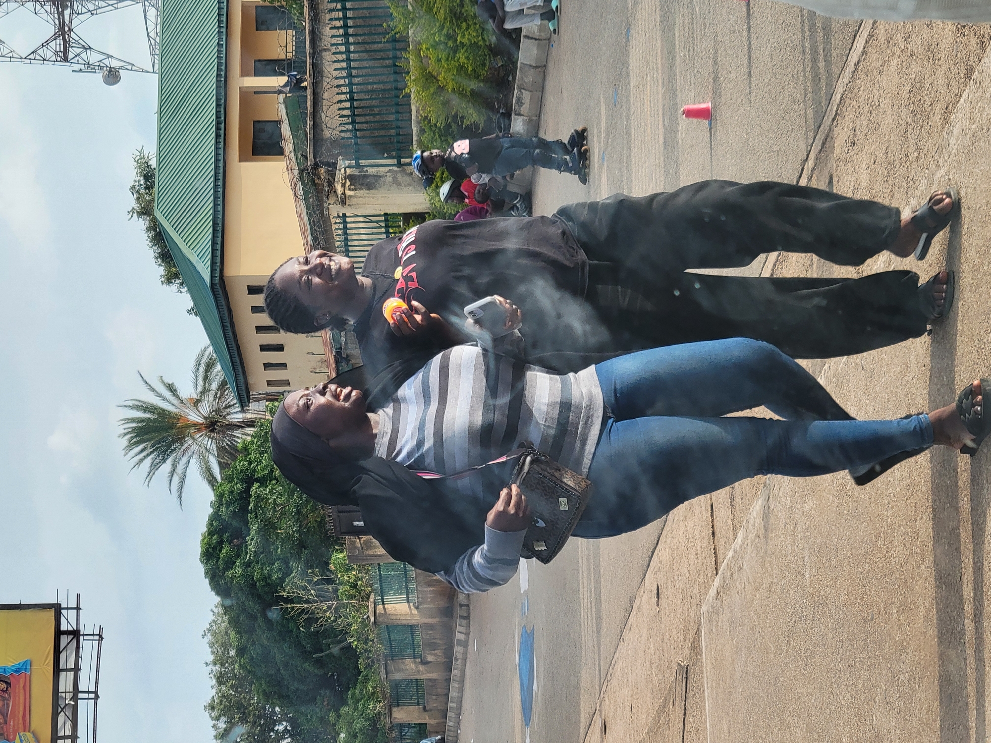 Two dark-skinned women are standing outdoors on a sunny day. One woman is wearing a striped shirt and blue jeans and carrying a small brown purse. The other woman is wearing all black. They are both smiling and looking upward. The woman in black is holding an orange object and a smartphone, with smoke or vapor visible near the phone. In the background, there are trees, a building with a green roof, a metal fence topped with barbed wire, and other people, including a roller skater. A red plastic cup is on the ground nearby. The image appears to be taken in Jos, Nigeria. A tall metal tower is visible in the background.