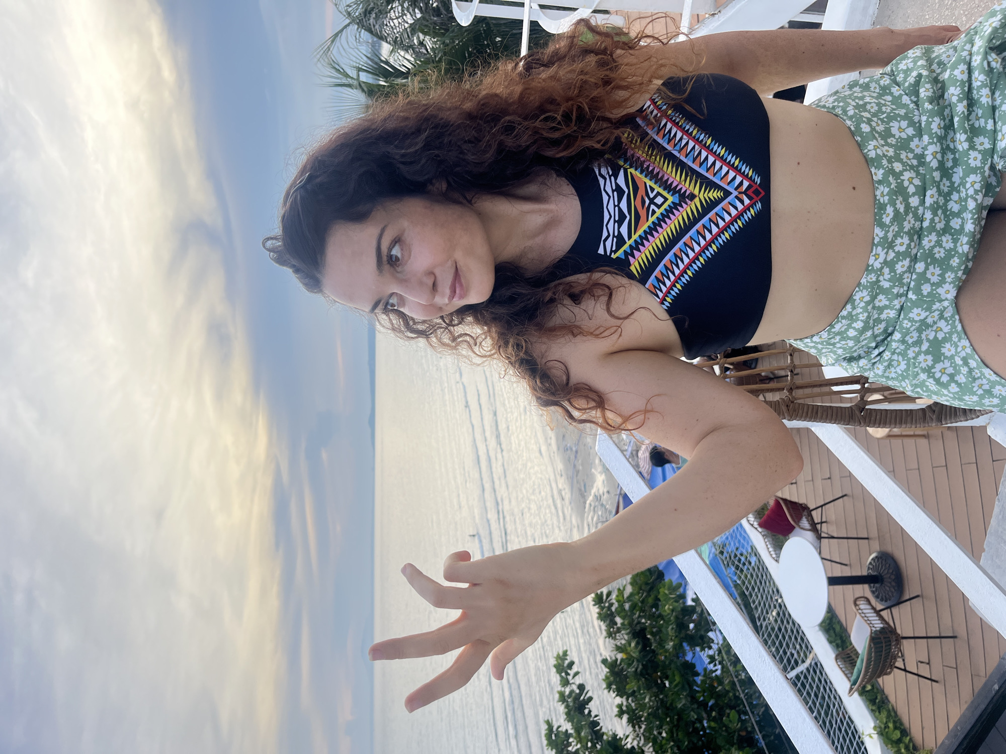 A woman with long, curly brown hair is pictured outdoors, likely in Mũi Né, Vietnam. She is wearing a black bikini top with a colorful geometric pattern and a light green skirt with a floral print. She is seated on a chair, leaning forward and looking towards the camera with a slight smile and raised eyebrows, suggesting a playful or curious expression. Her left hand is extended forward, with her fingers forming a gesture.
The background shows a body of water, likely the sea, with gentle waves and a pale, cloudy sky. The light suggests it is either early morning or late afternoon. There is a white railing in the foreground, indicating she is on a balcony or deck overlooking the water. In the background, part of an outdoor seating area with tables and chairs is visible. Green foliage, possibly palm trees or other tropical plants, can be seen on the right side of the image. The overall atmosphere is relaxed and scenic. No visible text is present in the image.