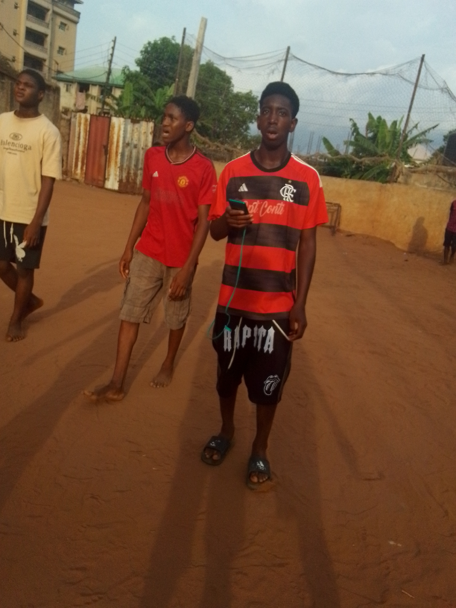 Three adolescent males stand on a brown, sandy ground with a building and vegetation in the background. The male on the left wears a light-colored t-shirt and dark shorts and is barefoot. The second male wears a red soccer jersey and tan shorts, also barefoot. The third male wears a red and black striped jersey, black shorts with white lettering, and blue slides. He holds a black cell phone. The location appears to be outdoors in the city of Unknown, Unknown. A metal fence and netting are visible in the background.