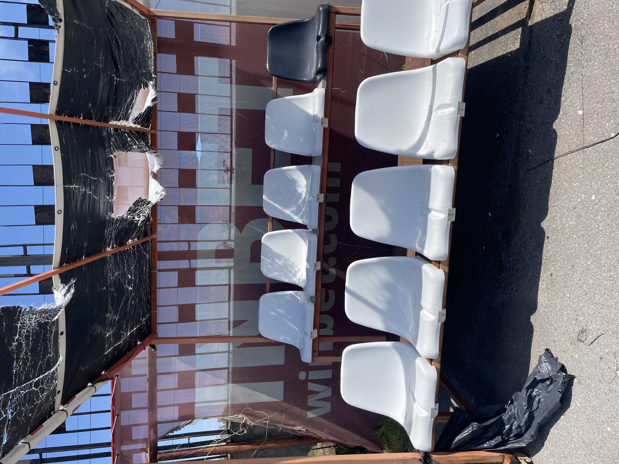 The image captures a section of an outdoor seating area, likely a temporary grandstand or a dilapidated sports venue, under bright daylight. Rows of empty plastic stadium seats are mounted on a light-brown metal or wooden frame. The front row displays five white seats, while the row behind it has four white seats and one black seat at the top.
A heavily torn and frayed black fabric, possibly a shade net or tarpaulin, covers the upper left portion of the structure, allowing glimpses of a clear blue sky. Behind the seats, a translucent panel or screen features a large advertisement or sign. The white text "Willi" and "com" is clearly visible against a reddish-brown background, suggesting a brand name such as "Willcom" or "William". Reflections of the sky are also visible on this background panel.
The ground beneath the structure is paved asphalt or concrete, with distinct, elongated shadows indicating strong, direct sunlight, suggesting a midday or early afternoon setting. A crumpled black plastic bag lies on the ground near the base of the seating. No people or activities are present in the scene, which appears to be in a state of disrepair.