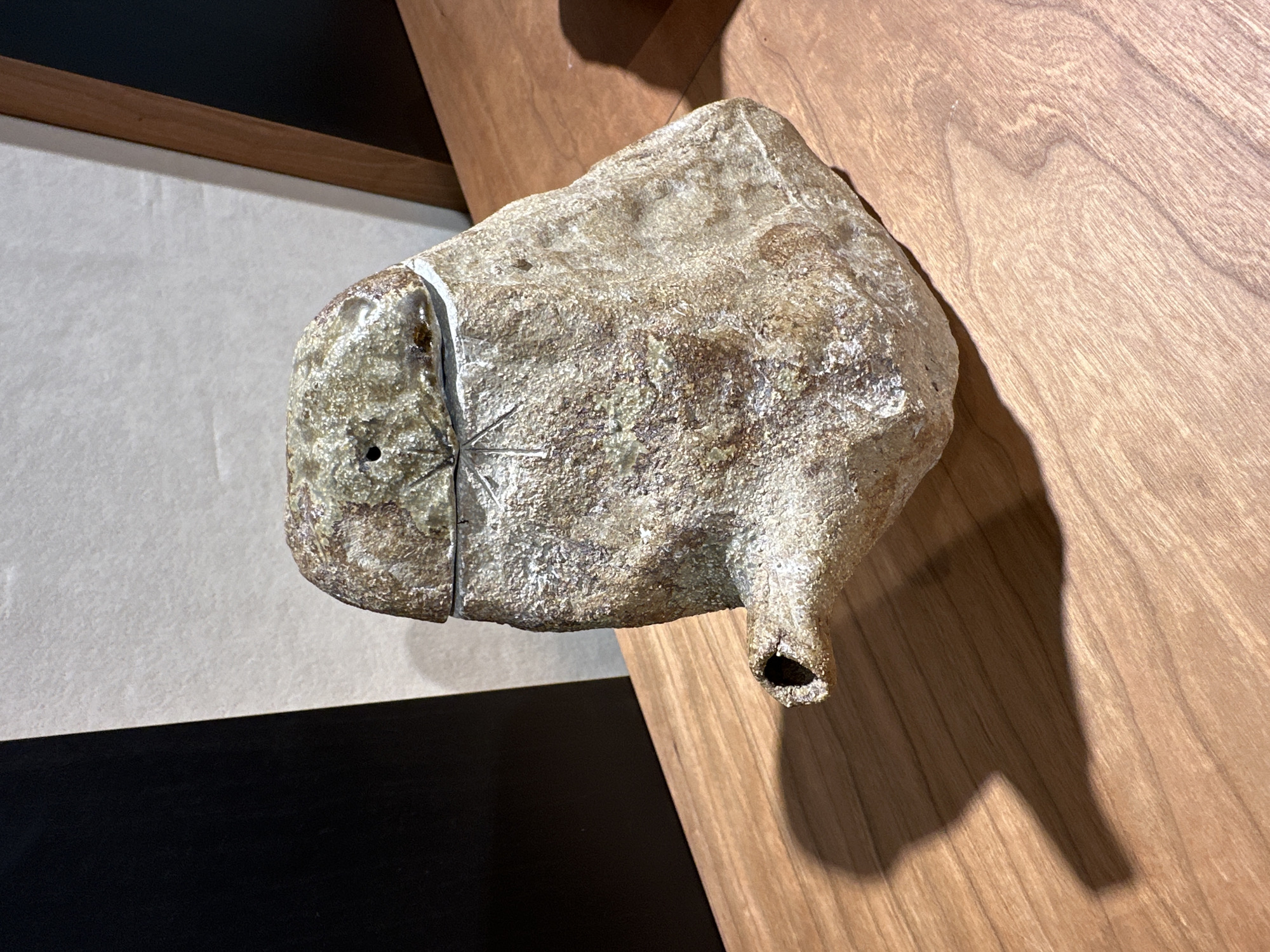 A unique, rough-textured object, possibly a ceramic sculpture or a natural stone artifact, is prominently displayed in an indoor setting. The object has an irregular, organic shape, appearing fuller and rounded on its left side and tapering to a short, hollow spout on its right. Its surface is mottled with varying shades of light brown, grey, and hints of green, giving it a coarse, granular, and weathered appearance.
On its left side, a thin vertical line or seam runs down, possibly indicating a join or a decorative groove. A small, dark, circular hole is visible on this part of the object. Near the center-left, a subtle star-like or asterisk-like engraving is etched into the surface. The hollow spout on the right has a dark opening, suggesting it could be a functional element or a design feature.
The object is positioned against a varied background. To its right, a warm-toned wooden panel or wall with distinct grain patterns serves as a backdrop. A strong light source, likely from the upper left, casts a clear, elongated shadow of the object onto this wooden surface. To the object's left, a light-colored, textured surface, possibly paper or a wall, is visible, with a contrasting dark black section below it. In the upper left corner, a dark brown or black structural element, perhaps a beam or frame, separates the light surface from another wooden section.
The scene suggests an exhibition or display in an art gallery, museum, or a well-curated interior. No people or visible text are present. This setting is located in London, United Kingdom.