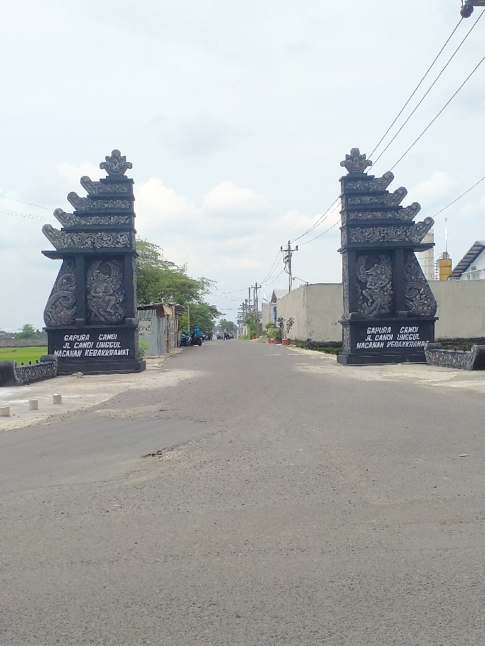 The image captures a straight-on view of a paved road flanked by two ornate *gapura*, or traditional Indonesian split gates, marking an entryway. The scene is set outdoors under a partly cloudy sky, suggesting daytime.
The two prominent *gapura* are dark in color, likely stone or concrete, and feature intricate carvings, including tiered roof structures and figures on their lower sections. On the base of the left gate, white text is clearly visible, reading: "GAPURA CANDI", "JL CANDI UNGGUL", and "MACANAN KEBAKKRAMAT". Similar text is partially visible on the right gate, with the OCR suggesting "JL CANDIN LINGGUL" and "NACANAN PEDAKKRANAJ", likely intended to be identical to the left gate. These texts, particularly "Kebakkramat," confirm the location context of Kecamatan Kebakkramat, Indonesia.
The asphalt road extends into the distance, showing some signs of wear such as cracks and minor potholes. Utility poles and overhead power lines run along both sides of the road. To the left, beyond the gate, there's lush green vegetation, possibly agricultural fields or dense trees, and a few small, simple buildings. Several motorcycles are parked further down the road on the left, with a few indistinct figures of people visible nearby. To the right, a long concrete wall borders the road, interspersed with potted plants. Further down this side, houses with various rooflines, including a distinct white one, can be seen. No specific event or activity is prominently featured; the image presents a snapshot of a typical day in this locale.
