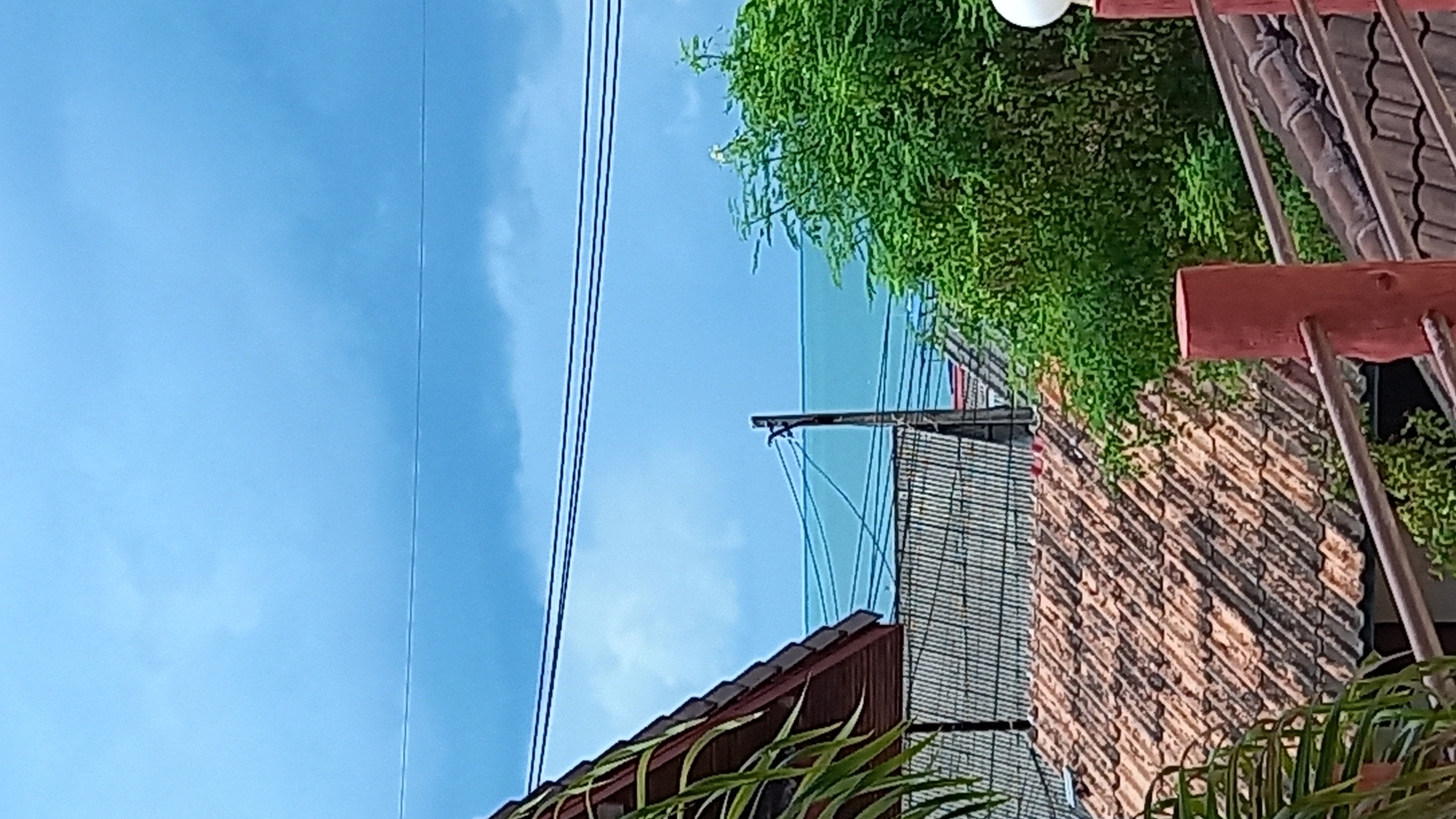 This is a close-up, eye-level, medium shot of a rooftop scene in Ko Pha-ngan District, Thailand, on a cloudy day. In the foreground, there is a terracotta tiled roof with some lighter colored tiles. A red wooden post with a rusted metal railing is visible on the right side of the image. Behind this, a lush green tree fills the right side of the frame. In the mid-ground, two more tiled roofs are visible, one with gray tiles and another with terracotta tiles. A wooden pole with electrical wires stretches across the frame horizontally. The background shows a body of water, likely the sea, with a bright turquoise hue, and a hazy blue sky with scattered clouds. There are no people or animals present in the image. The scene depicts a typical residential or resort area on the island.