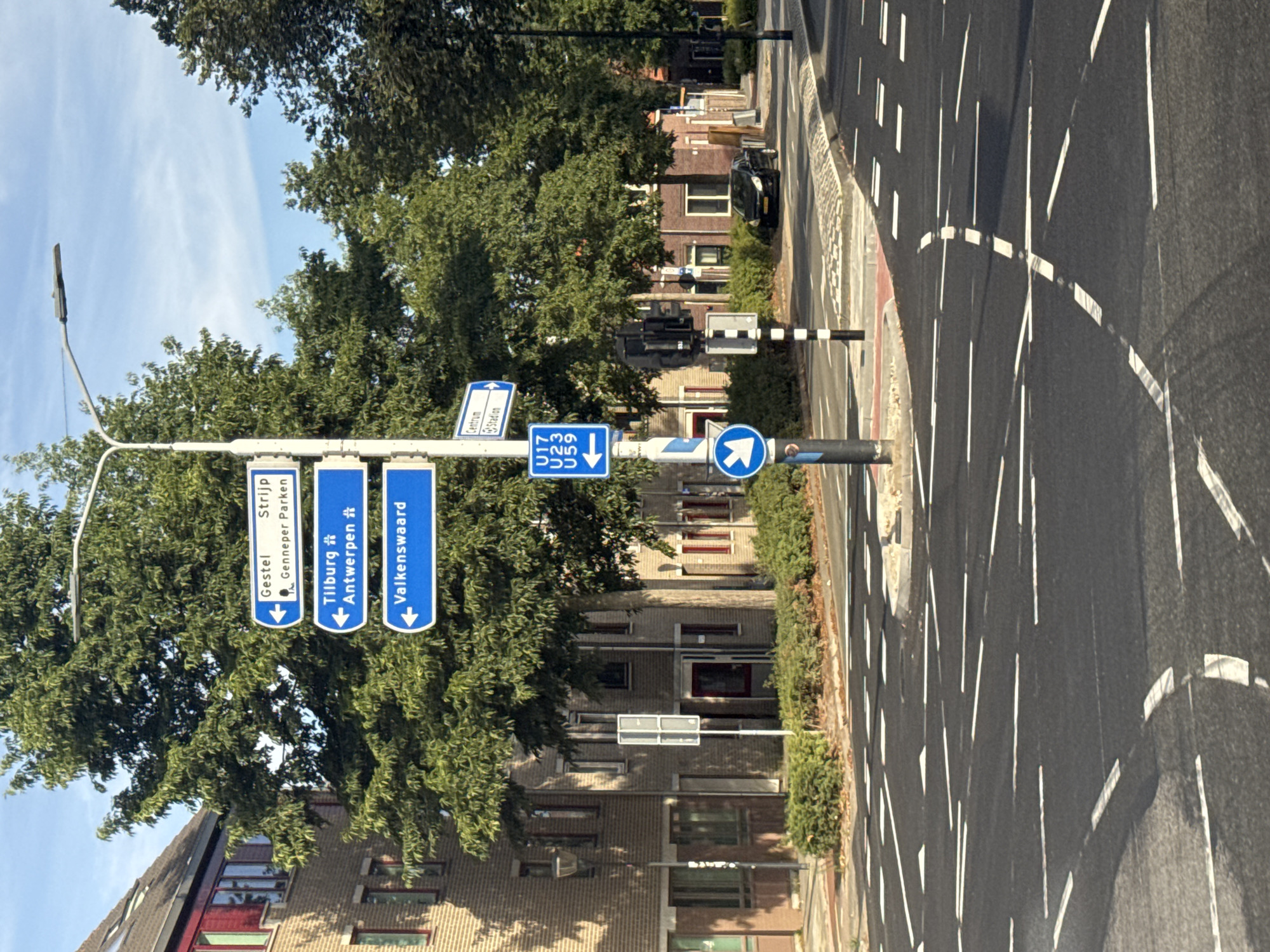 The image shows a street scene in Eindhoven, Netherlands, with a focus on a directional signpost. The signpost displays multiple blue signs with white text and arrows. The top sign indicates "Gestel Strijp" and "Gennep Parken" with an arrow pointing left. Below that, a sign reads "Tilburg" with an icon of a building and "Antwerpen" with a similar icon, both with arrows pointing left. The third sign points left and reads "Valkenswaard". A smaller blue sign on the lower right section of the post has white text "Centrum" and "Stadion" with arrows pointing right. Further down, another blue sign points left and displays "U17", "U23", and "U59". To the left of the signpost, a white pole with a blue circle and a white arrow pointing left indicates a mandatory direction. The environment includes a paved road with white lane markings, a traffic light, trees with green foliage, brick buildings, and a black car parked on the side of the road. The sky is blue with some clouds.