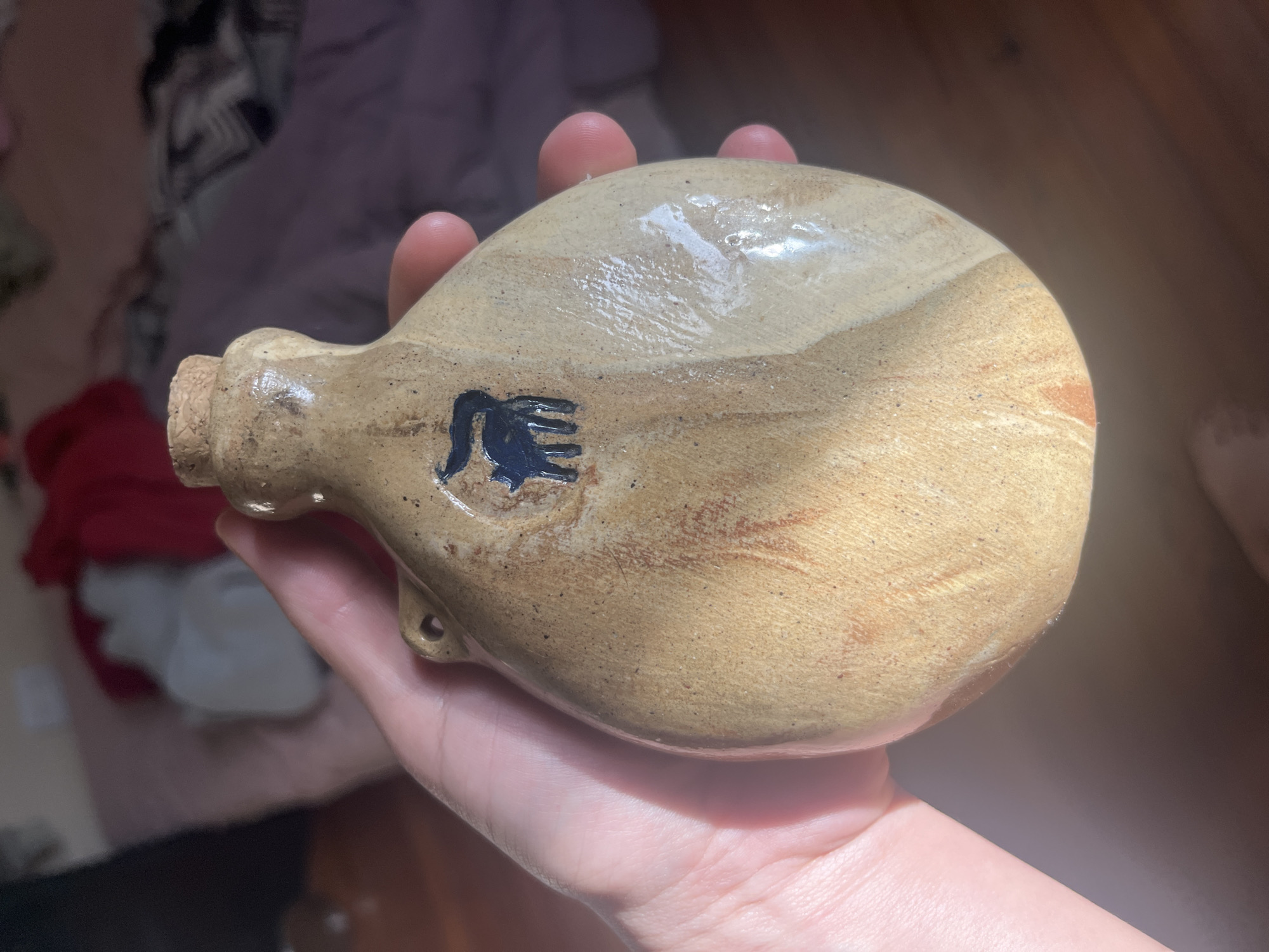 A person's hand holds a light-colored, possibly ceramic or stoneware, canteen-shaped flask in a close-up, indoor shot. The flask has an elongated, somewhat flattened oval body with a small neck and a cork stopper. Its surface is a mottled beige or tan, with areas appearing unglazed and others showing a subtle sheen, possibly from a clear glaze or liquid residue, creating highlights. Reddish-brown streaks and specks are visible across its surface, adding to its rustic appearance.
Near the base of the flask's neck, there are two small, unglazed holes, likely intended for a strap. A dark, abstract symbol, resembling a stylized animal figure or an emblem with flowing lines (perhaps a horse's mane and tail or a mythical creature), is prominently painted in black or dark blue on one side of the flask. The person's fingers gently curl around the flask, highlighting its ergonomic shape.
The background is softly blurred and out of focus, revealing indistinct shapes and colors, including hints of red, white, and a muted purple or grey, suggesting fabric or bedding. The lighting appears natural, casting soft shadows and emphasizing the texture and subtle variations in the flask's surface. No discernible text beyond the abstract symbol is visible. The scene takes place indoors, with no specific cues pointing to the provided location context of Buenos Aires, Argentina.