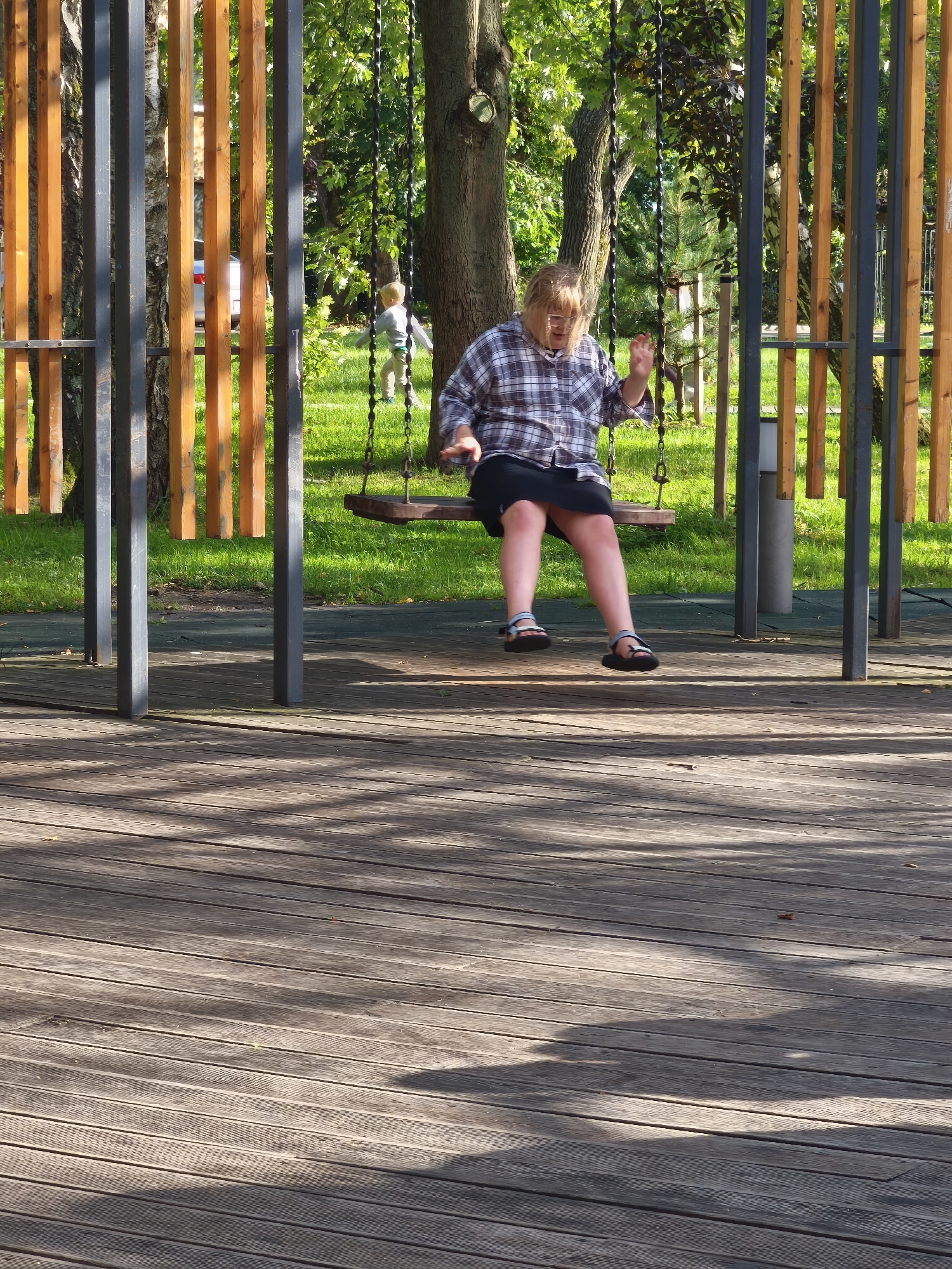 An outdoor scene features a woman on a swing set in Zelenogradsk, Russia. The woman, wearing a plaid shirt and dark skirt, is in mid-swing, her legs outstretched. She has light-colored hair and is wearing glasses. Behind her, a child in a light-colored shirt and pants stands on the grass. The swing set has dark metal supports and wooden slats, with a wooden seat. The ground in the foreground is a wooden deck. In the background, there are trees and green grass. Sunlight casts shadows on the wooden deck.