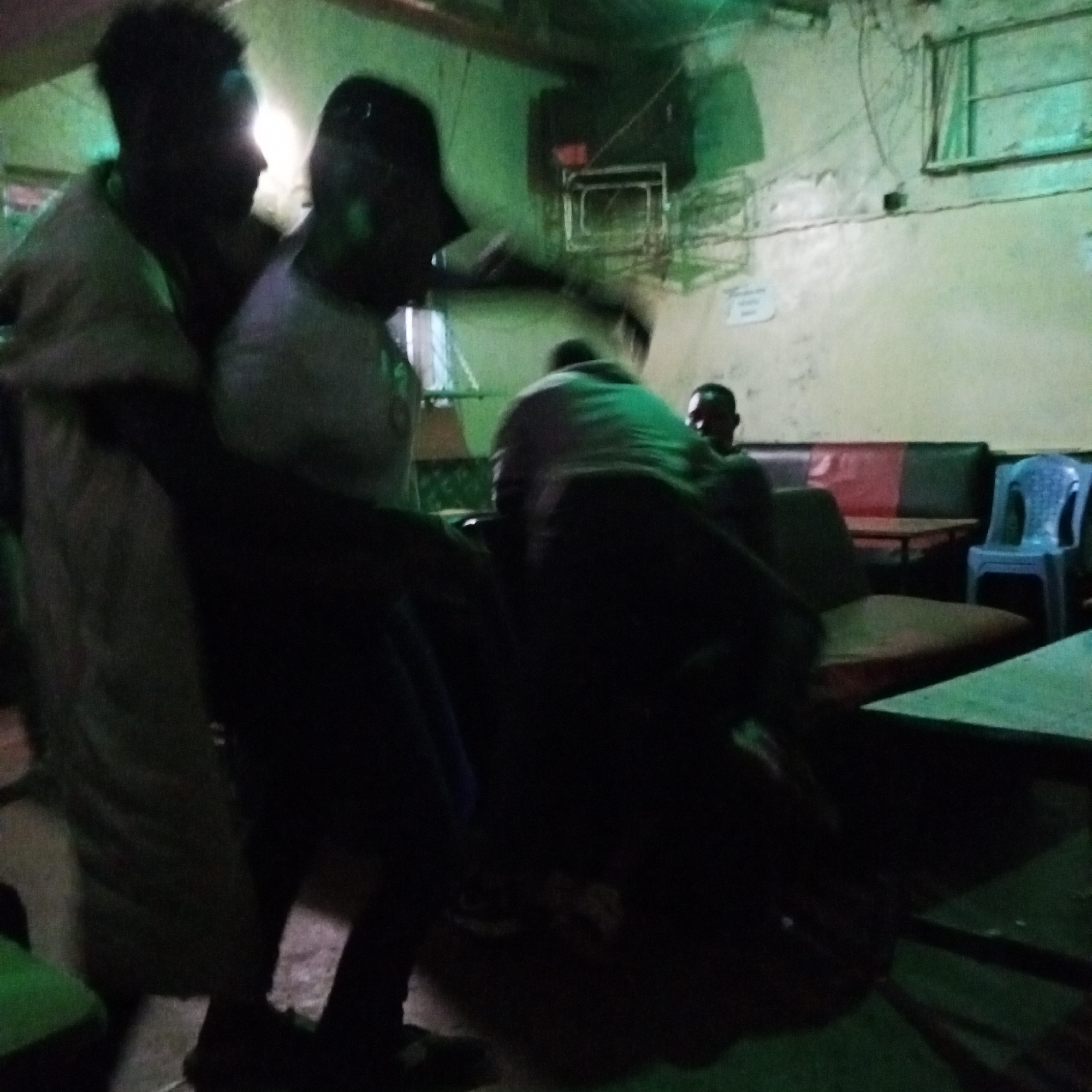 The image depicts an interior scene in what appears to be a bar or club in Borabu Chitago ward, Kenya. Two men are standing, one in a light-colored robe and the other in a t-shirt and cap. The man in the t-shirt is gesturing with his arm. Two other men are seated on a reddish-brown bench. There is a blue plastic chair to the right of the bench. A green-topped table is in the foreground. A television is mounted high on the wall. The lighting is dim and appears to be fluorescent.