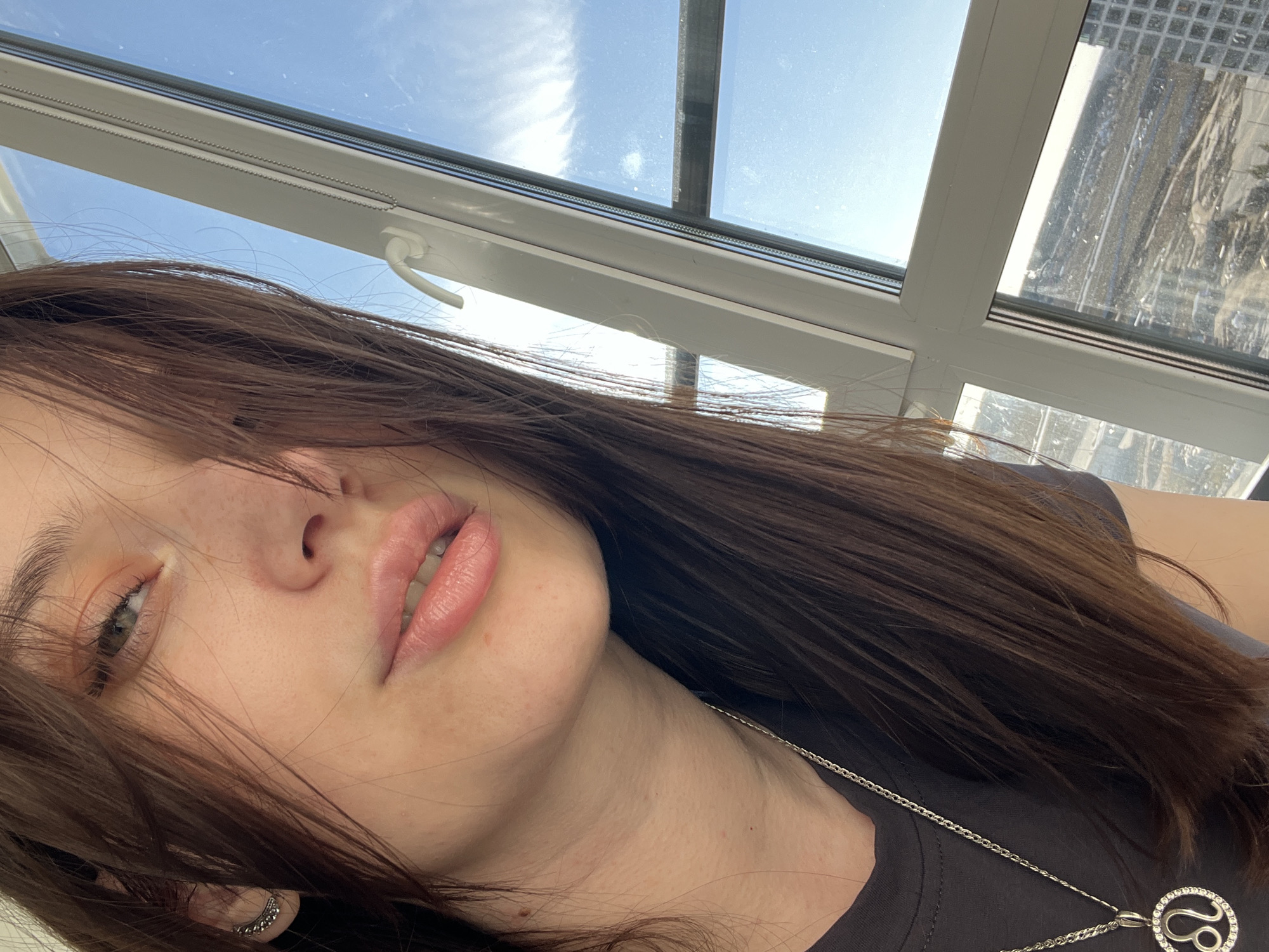 A close-up, tilted selfie captures a person with long, dark brown hair, partially obscuring their face. One eye, subtly made up, is visible, along with their nose and full, slightly parted lips. A silver earring or cuff adorns their visible ear. The person is wearing a dark t-shirt and a silver chain necklace with a circular pendant. The pendant features the astrological symbol for Leo, embellished with small, sparkling stones.
The background reveals a white-framed window, likely a skylight or an angled window, allowing bright natural light into the scene. Through the window, a clear blue sky with scattered white clouds is visible. Reflections on the glass indicate a sunny day. Beyond the window, a blurred urban or industrial landscape can be seen, characterized by distant structures with grid patterns, and what appears to be light-colored, possibly snow-covered, surfaces, suggesting a cold climate typical of Tyumen, Russia, the provided location context. The overall mood is relaxed and illuminated by natural daylight. No discernible text is present other than the zodiac symbol on the necklace.