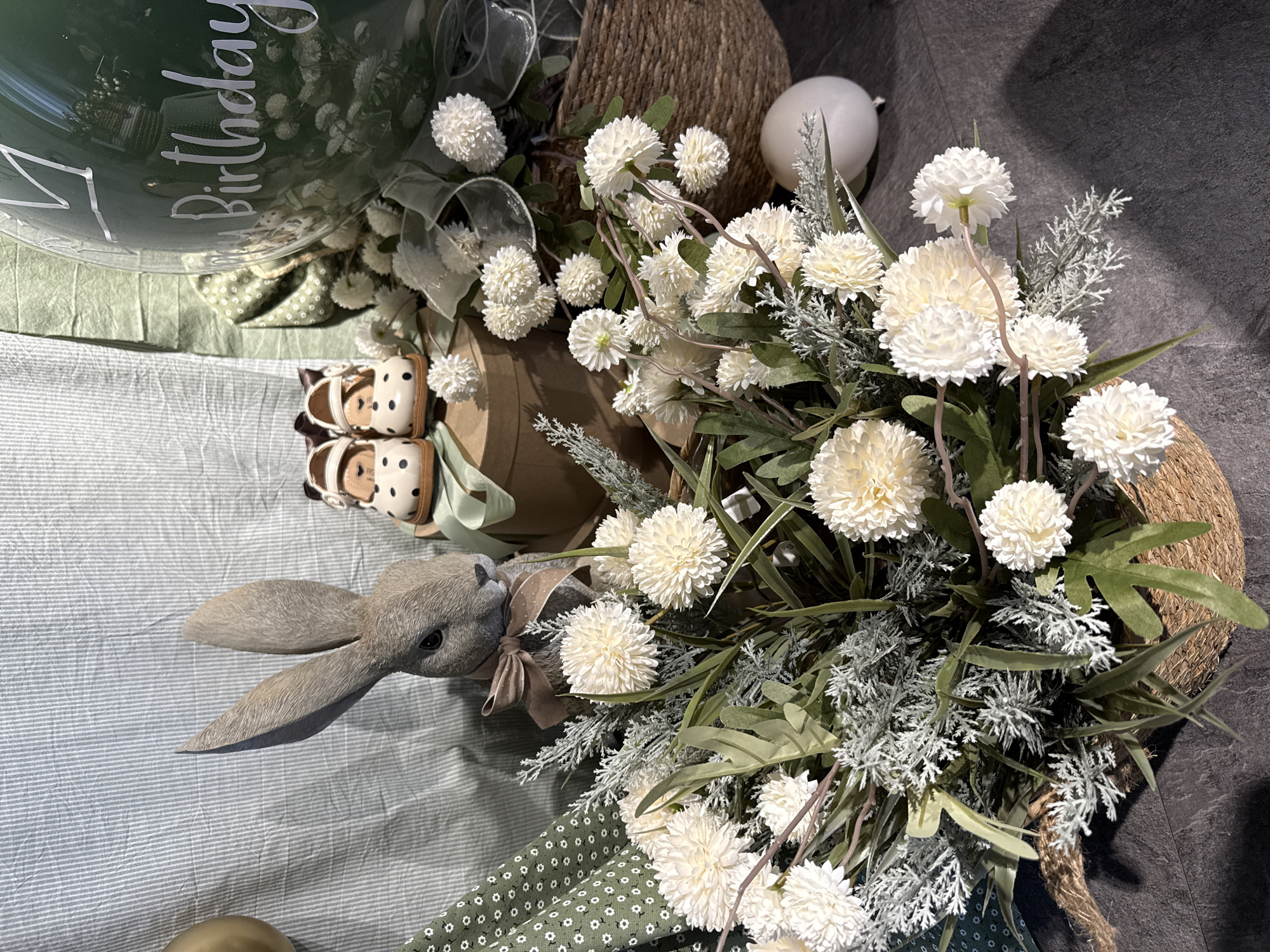 A flat lay of birthday decorations for a baby is shown. The primary subjects are a large green balloon with "Birthday" written in white script, a stuffed grey bunny with large ears, and a bouquet of artificial white flowers. The balloon is partially visible at the top left of the image. The bunny is positioned on the left side of the frame, lying on its side with its ears spread out. It has a brown bow tied around its neck. Next to the bunny are a pair of baby shoes with a polka dot pattern. The white flowers are arranged in a woven basket, filling the right side of the image and extending towards the center. The background consists of a textured surface that appears to be a floor or table, with some fabric elements and a neutral grey surface. The overall impression is a whimsical and celebratory setup, likely for a baby's first birthday. The lighting is soft and even, suggesting an indoor setting. There are no discernible cues about the time of day or weather, and the location context of Votkinsk, Russia is not visually evident. The scene evokes a sense of joy and anticipation.
