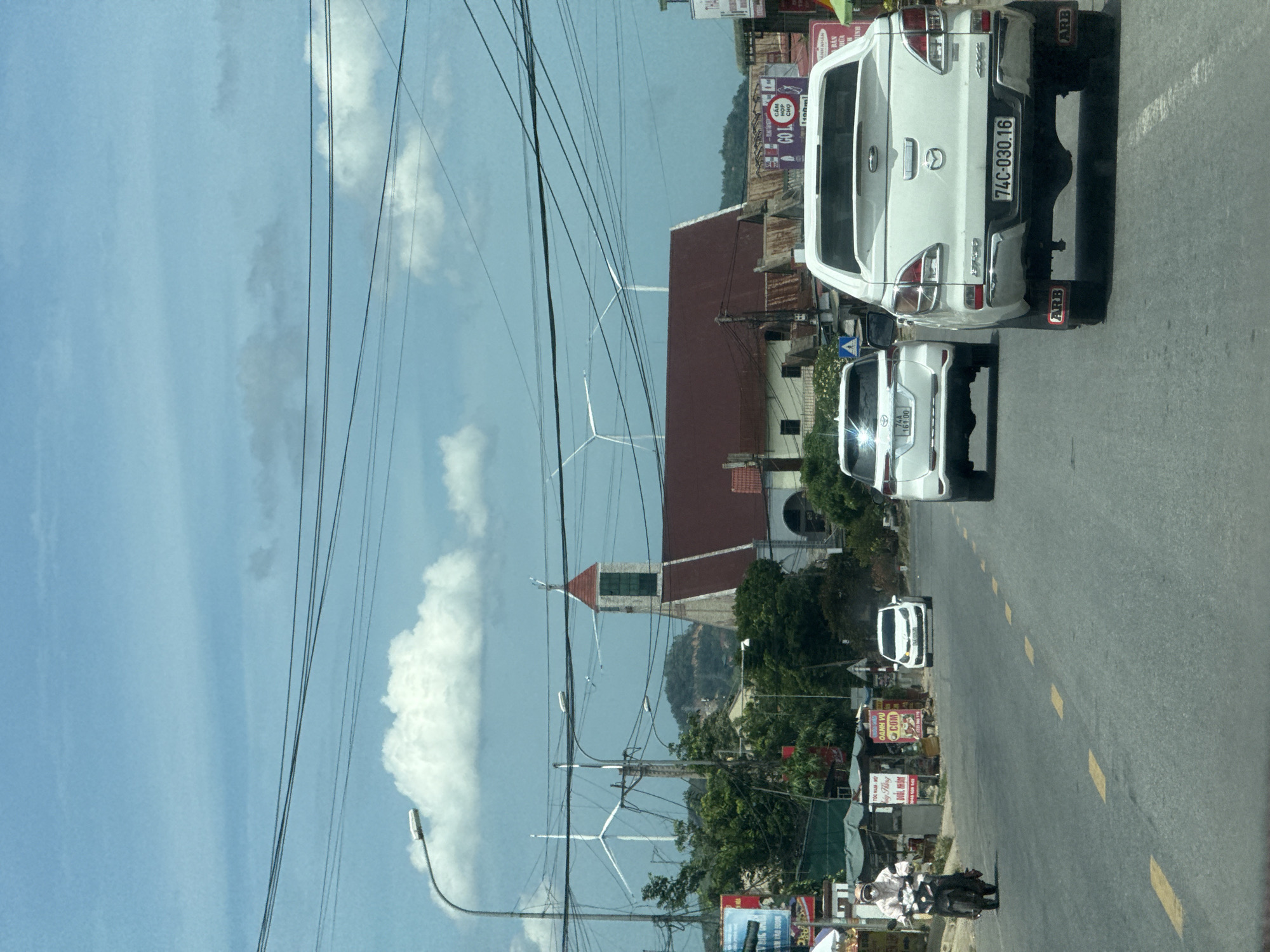 The image depicts a roadside scene in Tân Lập Commune, Vietnam, characterized by a bright, clear sky and scattered clouds, suggesting it is daytime. The road is paved and marked with yellow lines, indicating it's a thoroughfare. Several vehicles are visible, including two white pickup trucks and a white car. The first pickup truck, in the foreground, has a license plate reading "74C-030.16" and "ARB" decals on its mud flaps. Behind it, another white car displays a license plate "74A 161.00". A motorcycle with a rider is also present, partially obscured.
The surroundings are a mix of urban and natural elements. Buildings, some with red tiled roofs, line the road. Signs with Vietnamese text, such as "TÓC NAM - NỮ" (Men's - Women's Hair) and "OANH VÙ COM" (a food-related phrase), are visible, providing local context. In the background, lush green trees and a hillside can be seen. Notably, several wind turbines are present in the middle ground, suggesting a rural or semi-rural area with renewable energy infrastructure. Power lines crisscross the sky, a common sight in many regions. The overall impression is a bustling but somewhat rural roadside environment in Vietnam.