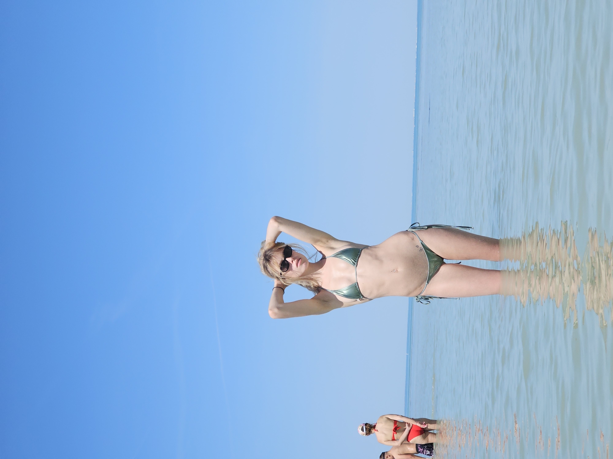 A woman in a green bikini and sunglasses stands in shallow water with her hands behind her head. The clear blue sky extends over the left half of the image, while the calm, shallow water fills the right half. Two people are visible in the water in the lower portion of the image, partially obscured by the reflection on the water's surface. The location is La Brée-les-Bains, France, as indicated by image metadata.