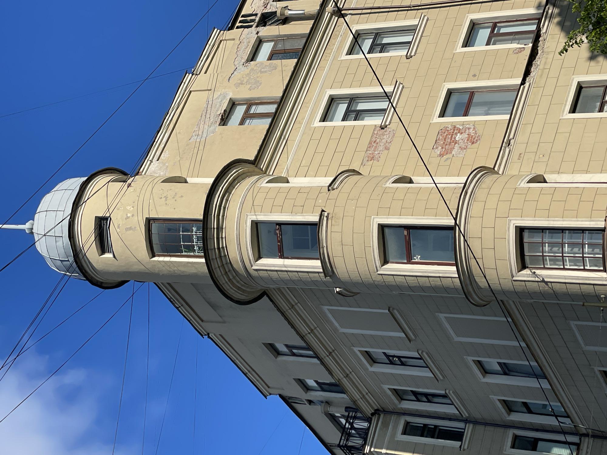 The image captures a building in Saint Petersburg, Russia. It showcases a beige building with several windows, some of which reveal interior details, and a cylindrical tower-like structure topped with a gray, dome-shaped roof. Black wires are crisscrossing in the sky near the tower. The building's facade shows signs of wear and tear, with exposed brick and peeling paint. The sky is blue with some clouds.