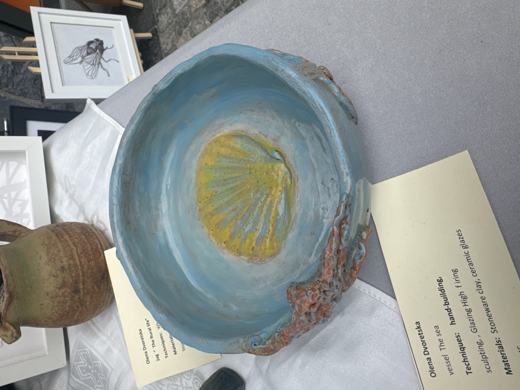 A ceramic bowl, likely a piece of art for sale, is prominently displayed on a table covered with a grey tablecloth. The bowl is blue on the outside with a textured, reddish-brown rim, and the inside features a yellow and blue swirling pattern resembling a seashell. Beside the bowl, a light yellow card provides information about the artwork, listing the artist as Olena Dvoretska, the artwork title as "The sea" (for a vessel), and detailing techniques such as hand-building, sculpting, and glazing. The materials are identified as stoneware clay and ceramic glazes. In the background, a rustic jug is visible, along with framed artwork, one depicting an insect. This scene appears to be at an outdoor market or craft fair, potentially within the city of Kilkenny, Ireland, given the typical setup for such events in historical Irish cities.