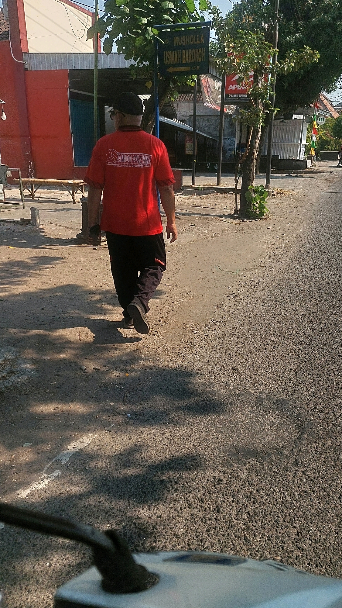 The image captures a street scene in Mangunsuman, Indonesia, likely during the day, indicated by the shadows cast by trees and buildings. In the foreground, the rear of a person walking away from the camera is visible. They are wearing a red t-shirt with a logo on the back that appears to be for "AMIGO CUP CAFE" and dark pants. The person is walking on a dirt and gravel path alongside an asphalt road. To the left of the path is a red building with a white trim and a light fixture hanging from the eaves. Further down the path, there are structures that suggest shops or small businesses. A green signpost with white text reading "MUSHOLLA USWAH BAROROH" and an arrow pointing to the right is visible on the left side of the road, along with another sign indicating "15 M". Trees line the area, with branches casting dappled shadows on the ground. In the far right background, colorful flags are visible, and a white building with a corrugated roof is present. The overall environment appears to be a semi-urban or rural setting. The perspective suggests the photo was taken from a low vantage point, possibly from a vehicle.