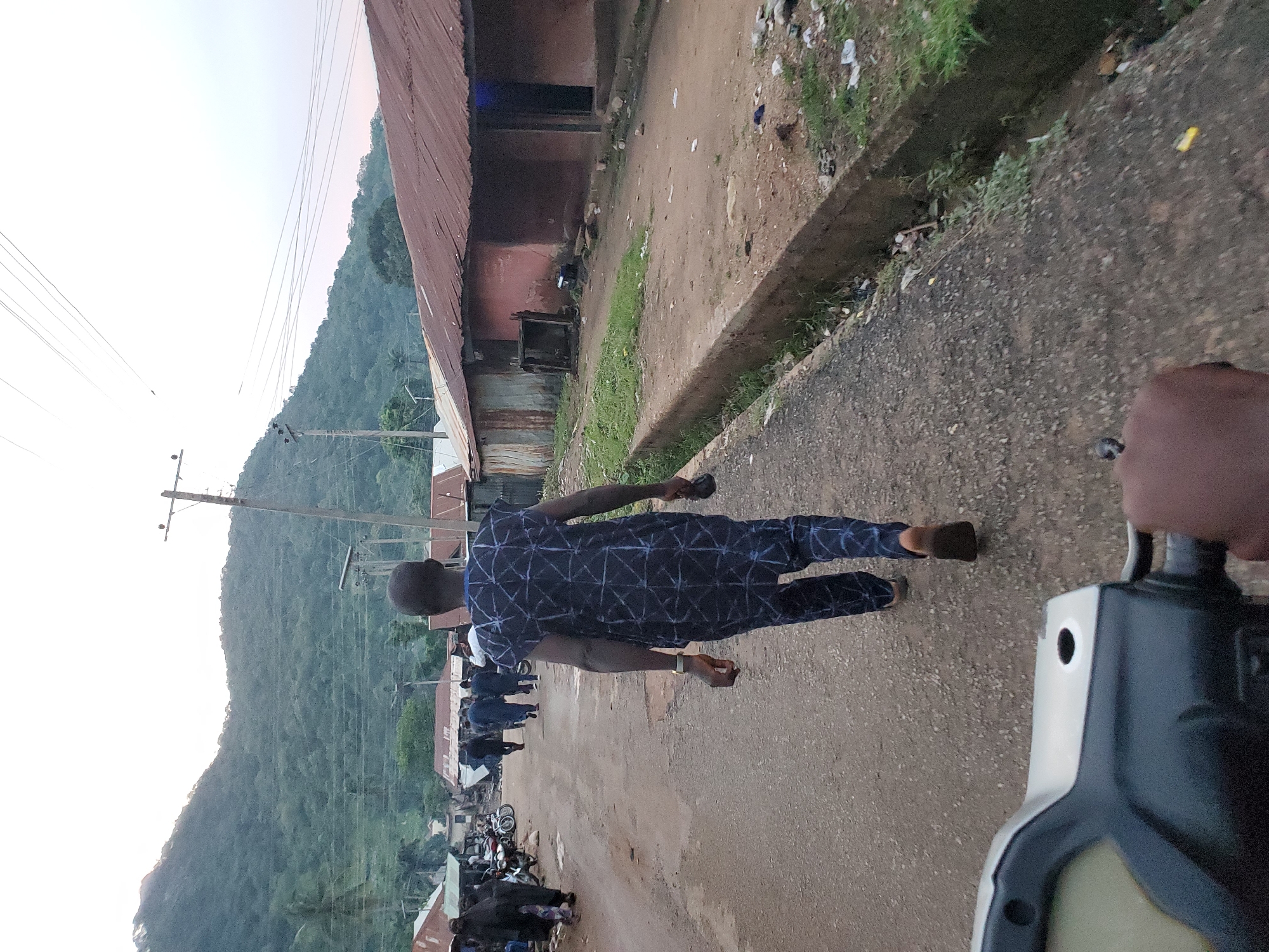 The image depicts a street scene in Mararaba, Nigeria, captured from a low perspective, likely from a scooter or motorcycle, with handlebars and a hand visible in the lower foreground. The lighting suggests either an overcast day or early morning/late afternoon.
In the immediate foreground, the back of a person is visible, walking away from the camera. This individual wears a dark blue garment adorned with a white geometric pattern. A gold-colored watch is present on their left wrist, and they hold a dark, indistinct object in their right hand. Their feet appear bare or in minimal footwear.
The central feature is an unpaved, dusty road exhibiting ruts and uneven sections. A concrete drainage ditch runs along the right side of this road, containing visible litter and some water. Adjacent to the ditch, a row of single-story buildings lines the street. These structures have visibly rusty corrugated metal roofs and walls painted in muted colors, including reddish-brown and grey. Some appear aged or in a state of disrepair.
Further down the road, multiple individuals are present. On the left side, near some parked motorcycles, a small group of people, one wearing extensive dark clothing, are walking towards the camera. Further ahead, another group of people, mostly in dark attire, is seen walking away from the camera.
The background is dominated by a large, heavily vegetated mountain range. Several wooden utility poles with multiple overhead wires span across the mid-ground, indicating a basic electrical or communication infrastructure. Green foliage and a tall palm tree are also visible on the left side, contributing to the semi-rural or peri-urban setting.
