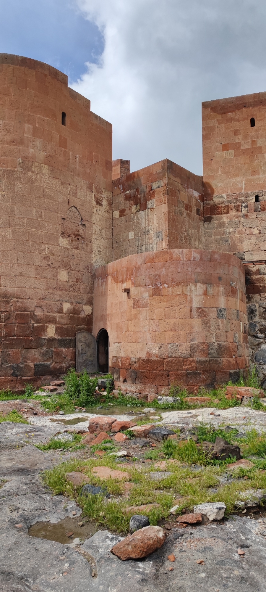 The image captures a stone fortress, likely located in the city of Dashtadem, Armenia, based on its architectural style. The fortress is constructed from reddish-brown stone blocks and features both square and cylindrical architectural elements. A weathered wooden door is visible, slightly ajar, providing a glimpse into the interior. The foreground consists of stone and brick fragments, scattered among patches of grass and puddles of water, suggesting a state of disrepair and potential recent rainfall. The sky is overcast, contributing to the overall muted lighting of the scene. No people are visible.