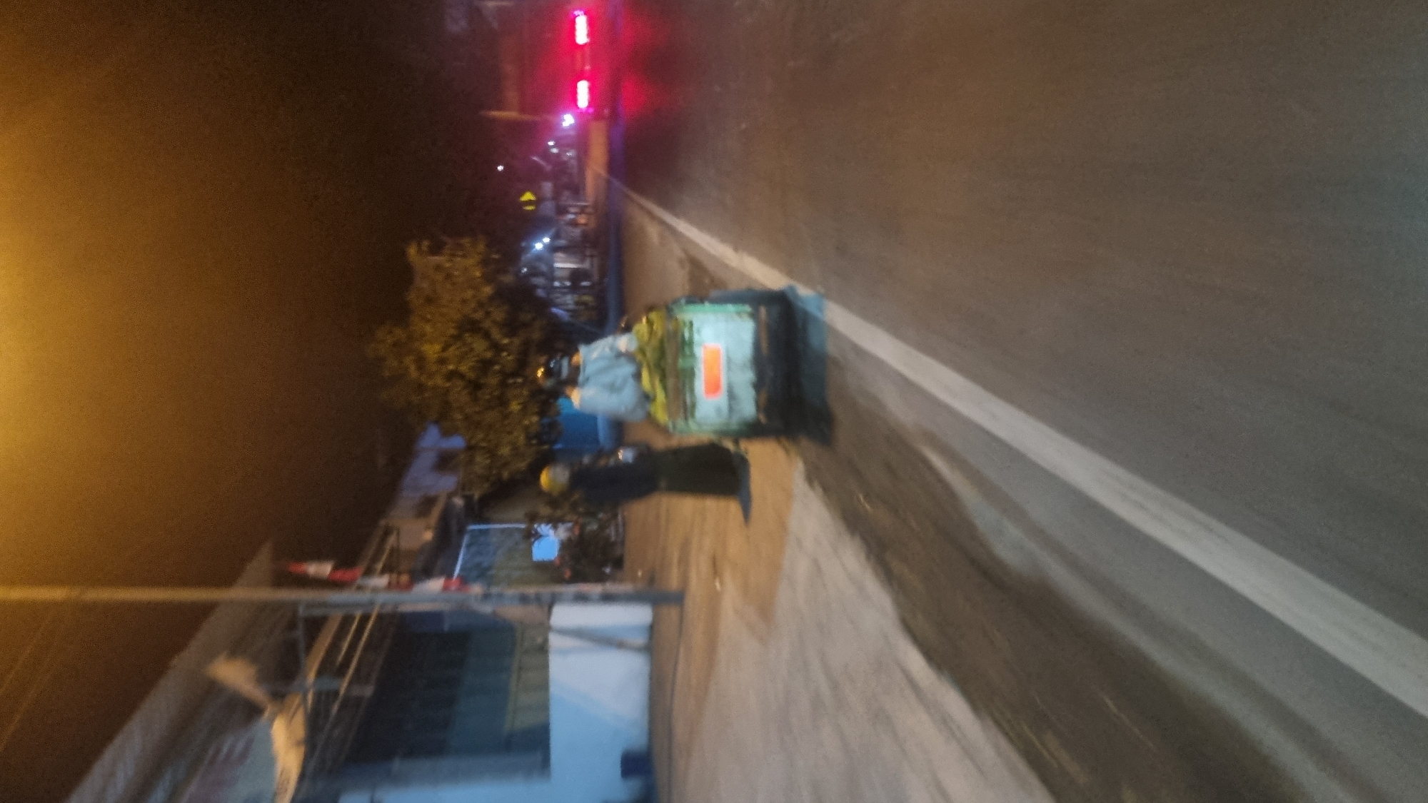 At night in Diwek, Indonesia, a person on a motorbike-pulled cart moves along a paved road. The cart is light green and appears to be carrying produce. The rider is wearing a light-colored jacket and a helmet. Beside the cart, on the sidewalk, another person dressed in dark clothing and a yellow headscarf stands. Buildings line the left side of the road, with streetlights illuminating the scene. In the distance, the taillights of a vehicle are visible. The sky is dark, suggesting late evening or night.