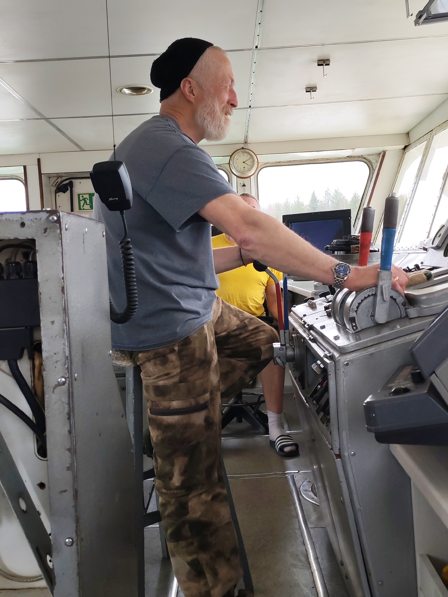 A man with a white beard and a black cap stands in what appears to be the control room of a vessel. He is wearing a gray t-shirt and camouflage pants. He is operating the controls, which include two levers with blue and red handles. Another person, wearing a yellow shirt, is seated behind him. The room has a clock on the wall, and there are windows showing a view of trees. The location is Depo, Russia.