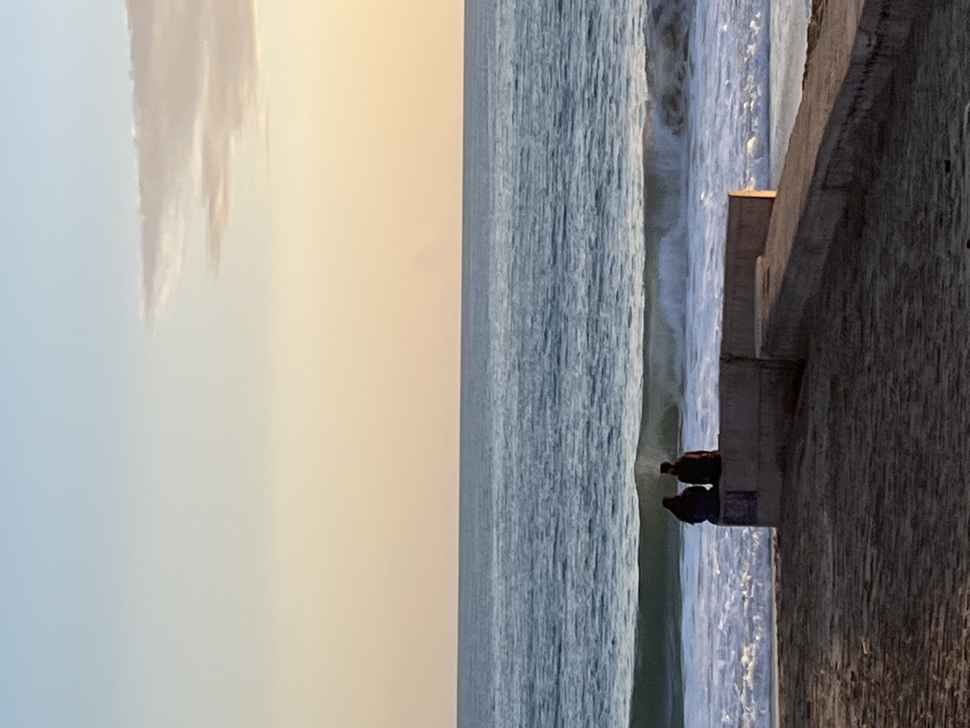 A vertical shot shows two people sitting on a concrete ledge overlooking the ocean at sunset. The sky transitions from light blue to a pale yellow and blends seamlessly into a white cloud formation. The ocean is directly adjacent to the ledge, with waves visible. The wall is dark and textured. The location is Alanya, Turkey.