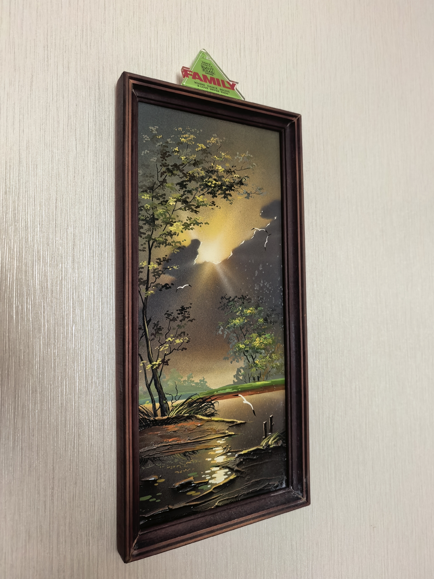 A framed painting of a sunset over a body of water is hanging on a textured wall. The painting depicts trees on the left side, with the sun setting behind them, casting a golden light. Birds are visible in the sky. The water in the foreground reflects the sunset colors. A green triangular sign with the word "FAMILY" in red is affixed to the top of the frame. The wall has a vertical striped texture and is a neutral color. The painting is oriented vertically.