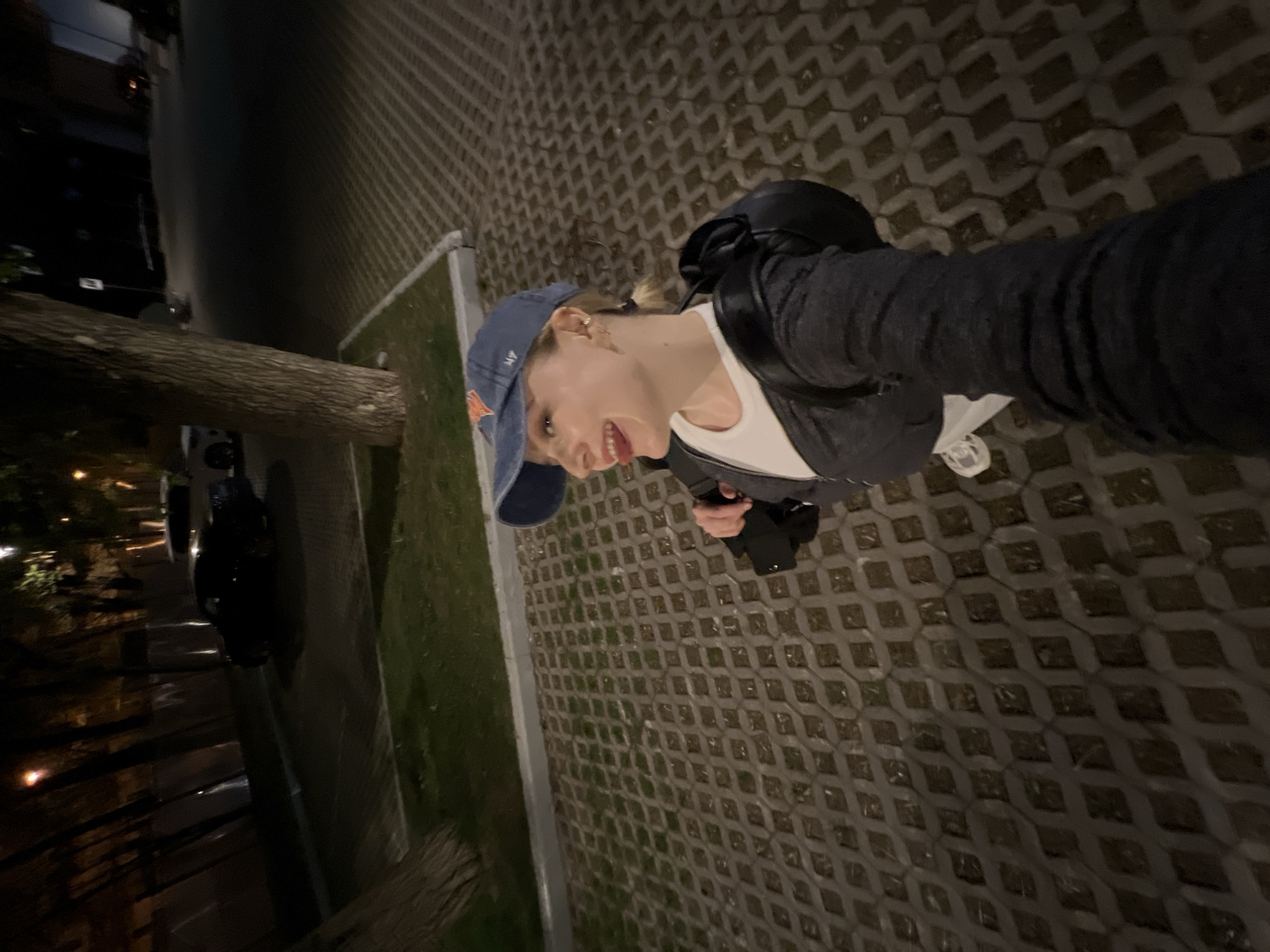 The image shows a person wearing a blue baseball cap, a white t-shirt, and a gray jacket, standing on a sidewalk in Khimki, Russia. The person is smiling and holding a phone in their hand, seemingly taking a selfie. A backpack is visible on their back. The sidewalk is paved with interlocking concrete pavers. To the left, there's a grassy strip next to a curb, with trees and streetlights in the background. In the distance, cars are parked along the street. The scene is captured at night.