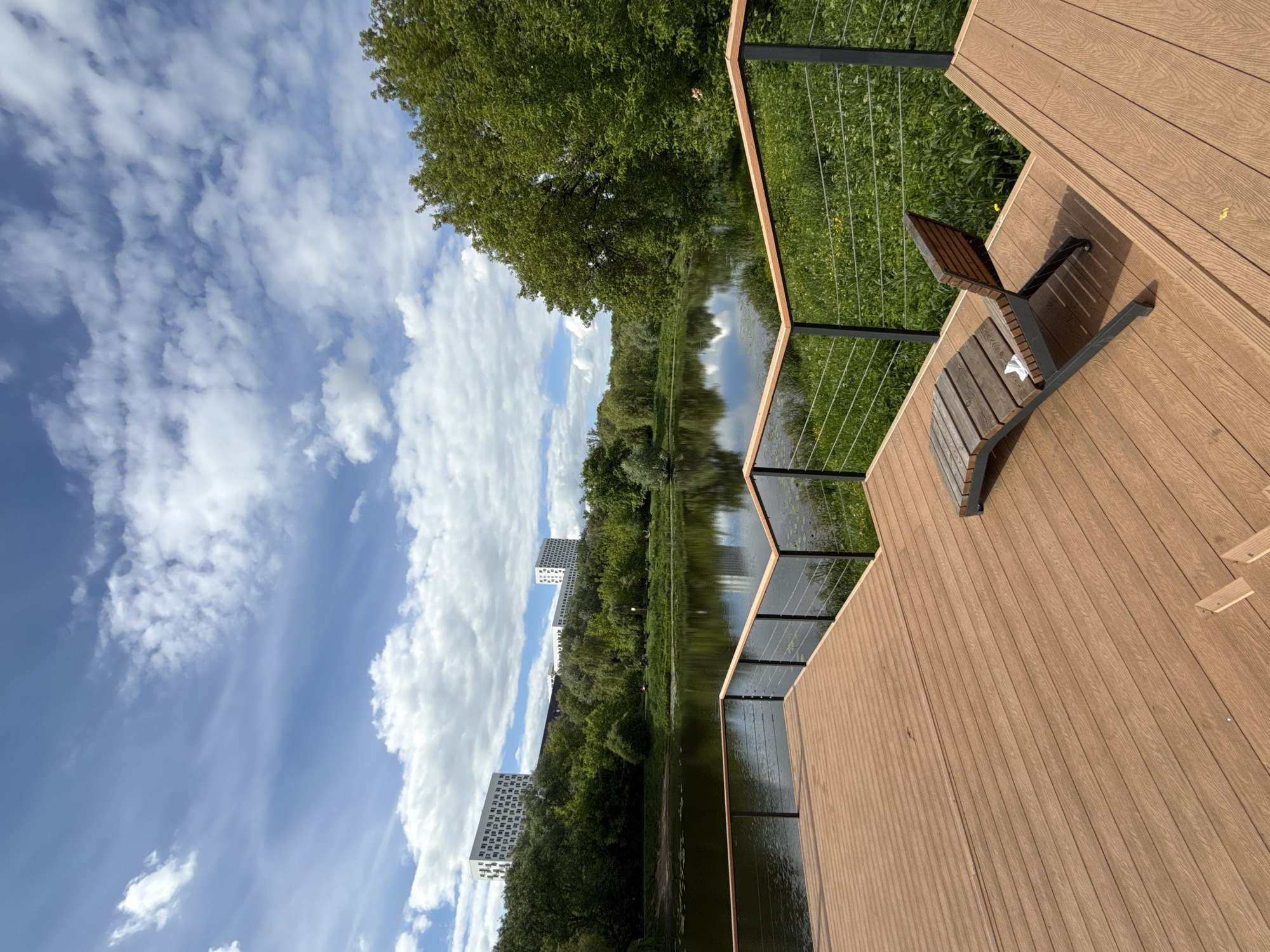 The scene shows a wooden deck overlooking a body of water, possibly a lake or a wide river. The deck features a railing with thin metal vertical supports, and a wooden bench sits on the deck. A blue sky with white clouds fills the upper left portion of the image. Trees line the opposite bank of the water, with some buildings visible in the distance. The photograph was taken during the day, likely in the city of Moscow, Russia.