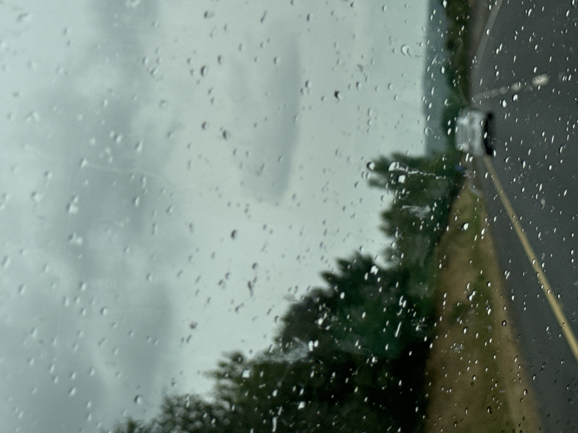 The image depicts a rainy day scene from inside a vehicle driving on a road. Water droplets are visible on the windshield, distorting the view of the environment outside. A grey, overcast sky dominates the upper portion of the image. On the left side, there are blurred green trees and foliage. The road ahead is asphalt with a yellow line marking the lane. A white car is visible in the distance, driving in the same direction. The background shows rolling hills. The scene is consistent with a view from a car traveling on a road in the countryside, possibly near Craan, Ireland, given the verdant landscape and the prevalence of rain. No people are clearly discernible in the image.